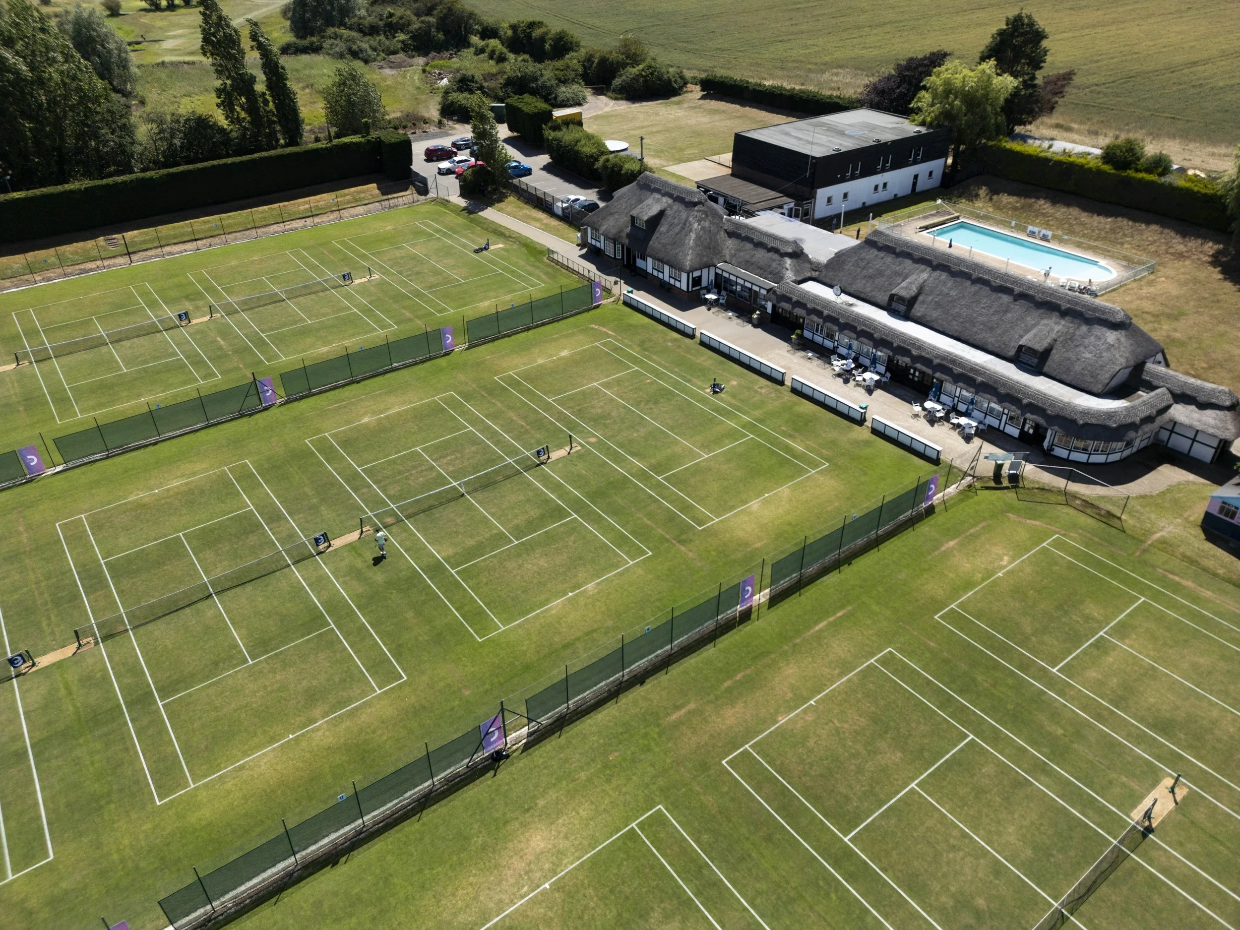 Aerial view of a tennis club with multiple courts, a main building with outdoor seating, a swimming pool, and parking lot, surrounded by greenery.