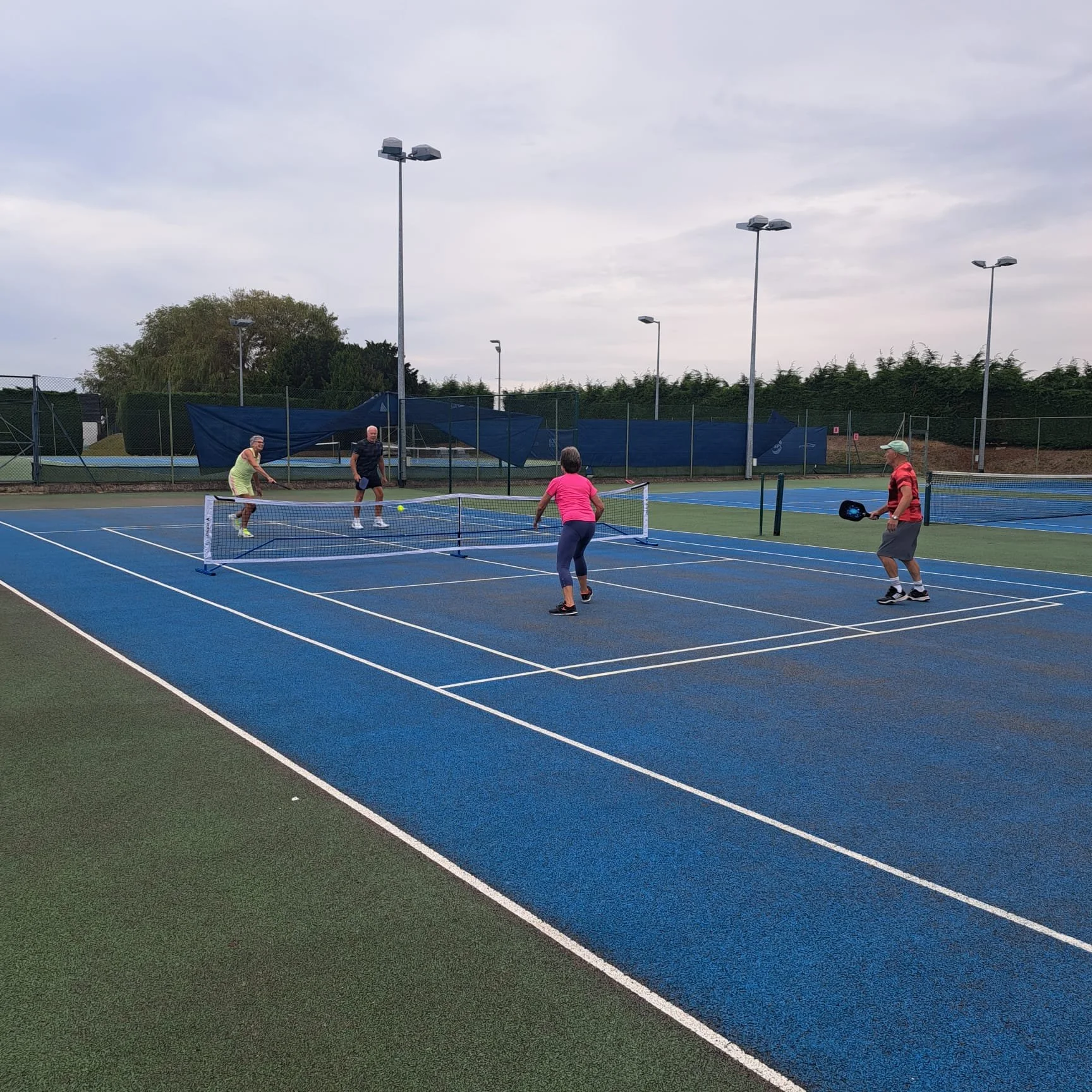 Four people playing pickleball on an outdoor court with blue surface, surrounded by a fence and tall light poles, under a cloudy sky.