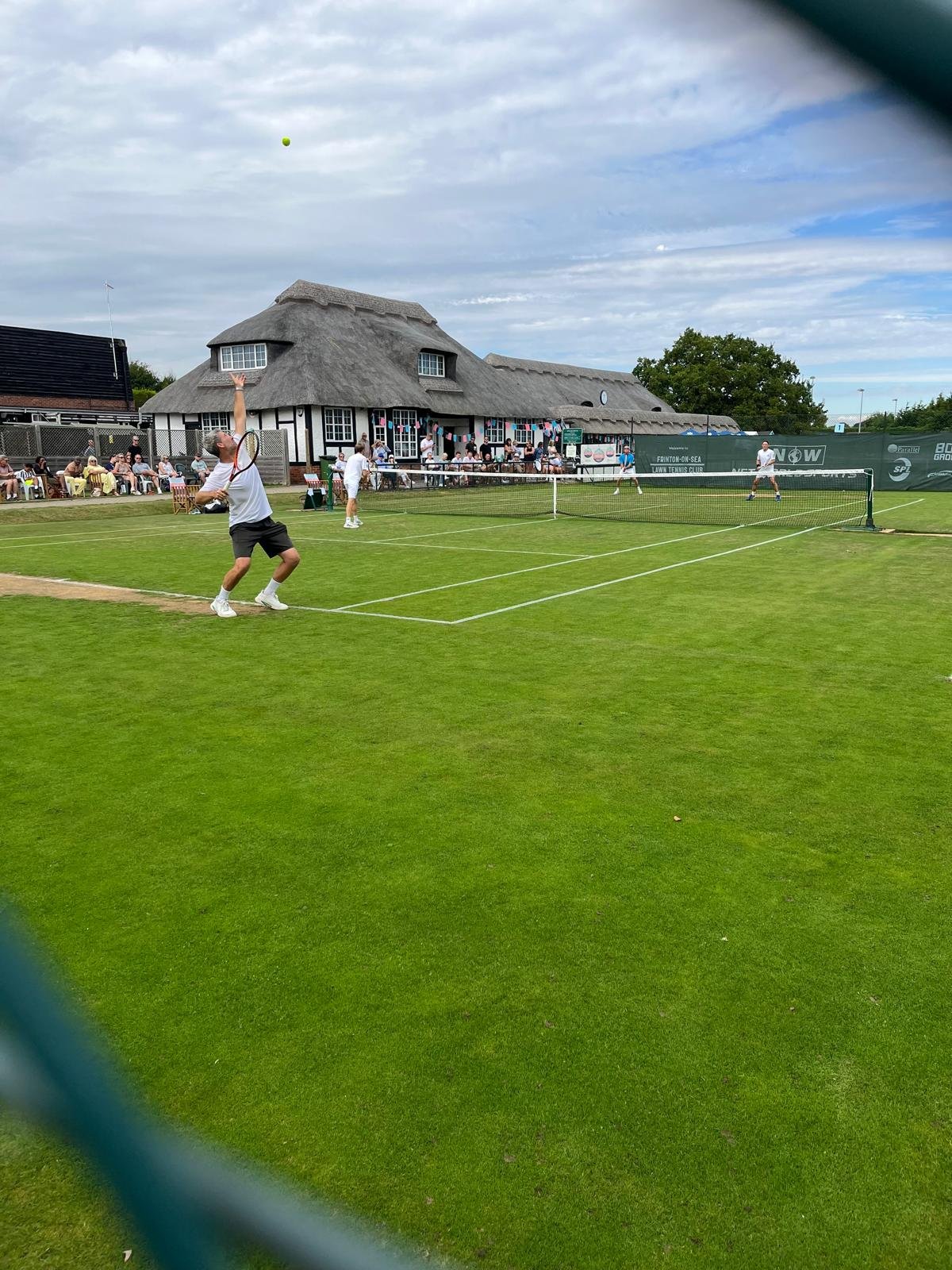 People playing pickleball on a grass court, with spectators sitting on benches and a thatched-roof building in the background.