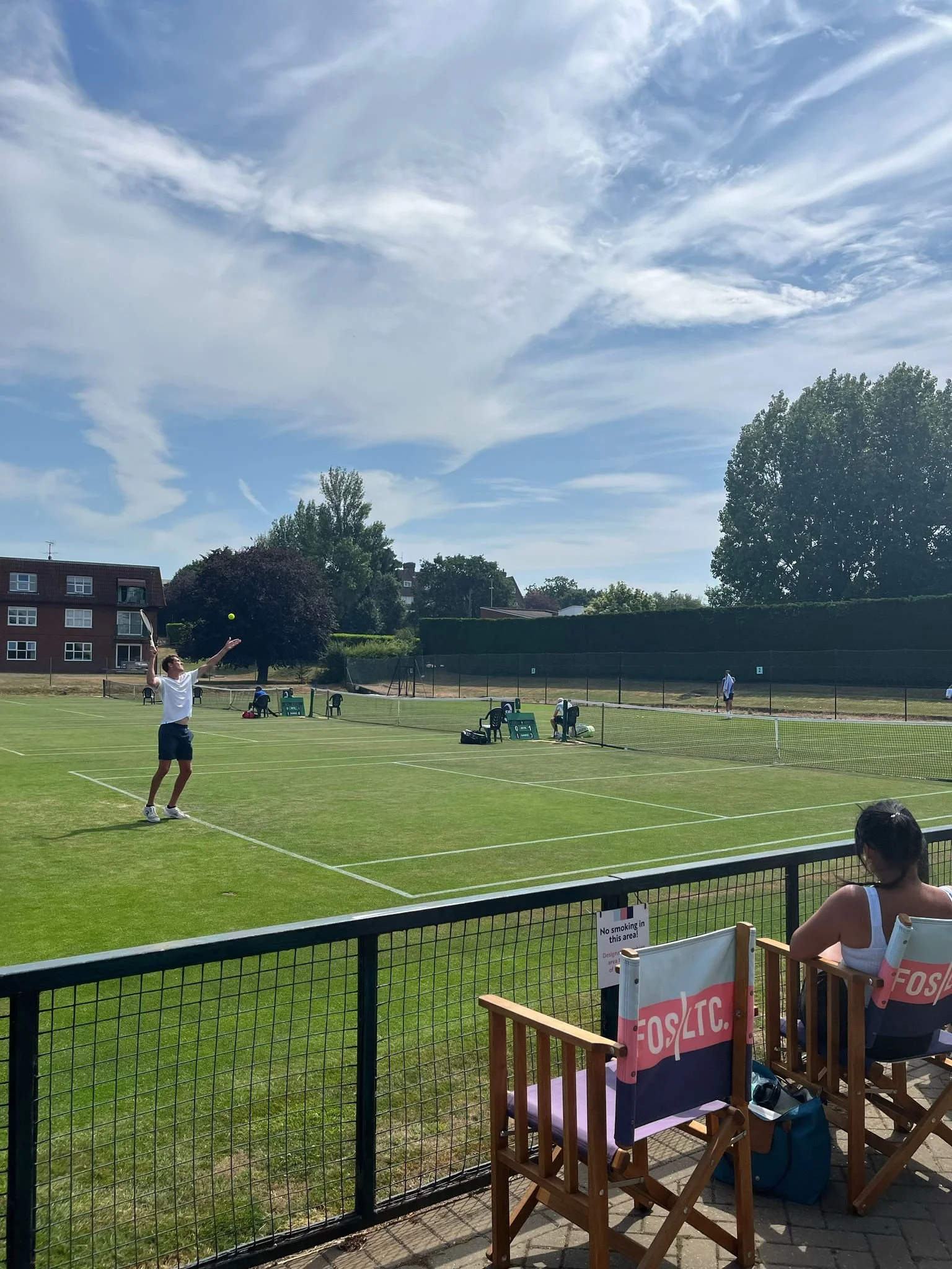A person playing tennis on a grass court, with spectators seated in chairs in the foreground. There is a woman sitting in one of the chairs, watching the game under a partly cloudy sky.