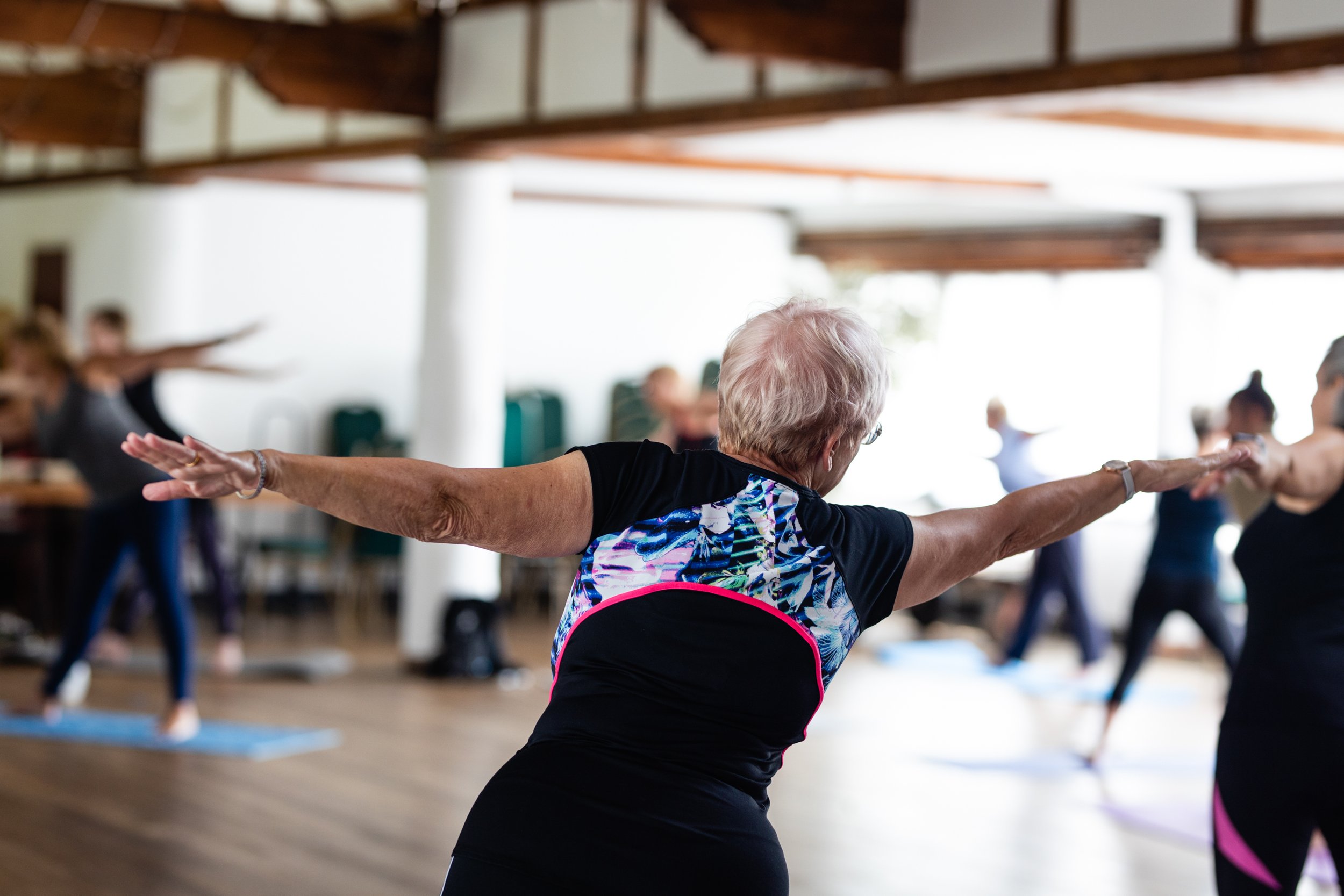 Senior woman participating in a yoga class, stretching with arms extended, in a spacious, well-lit studio with other participants in the background.