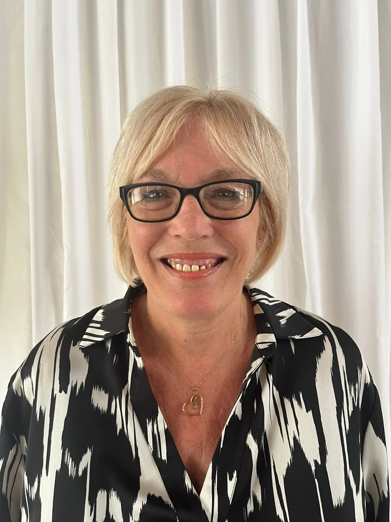 A smiling woman with blonde hair, wearing black-rimmed glasses, a patterned black and white blouse, and a necklace with a heart-shaped pendant, standing in front of white curtains.