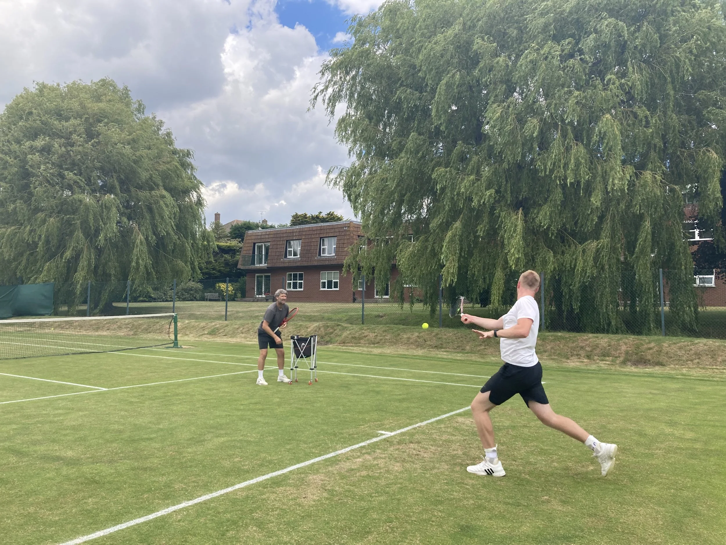 Two men playing tennis on a grass court in a park with large trees and a building in the background.