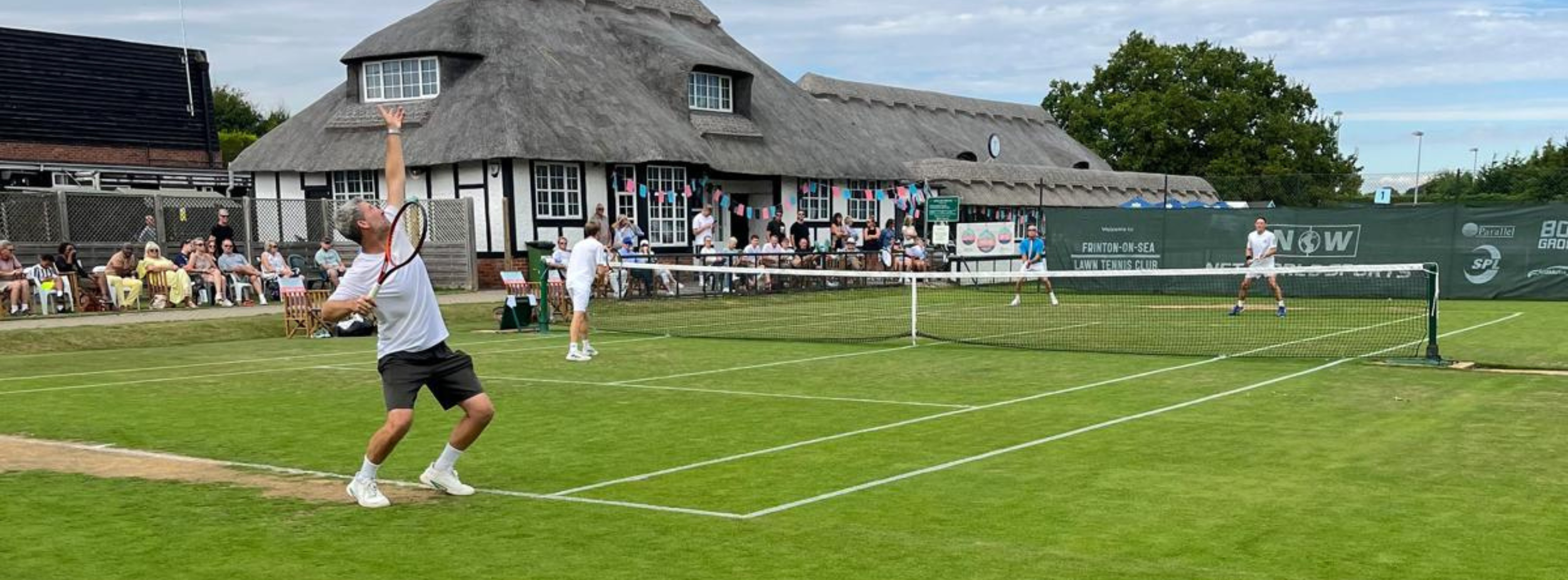 People playing pickleball on a grassy court with spectators watching from the sidelines, in front of a large thatched-roof building with umbrellas and colorful bunting.