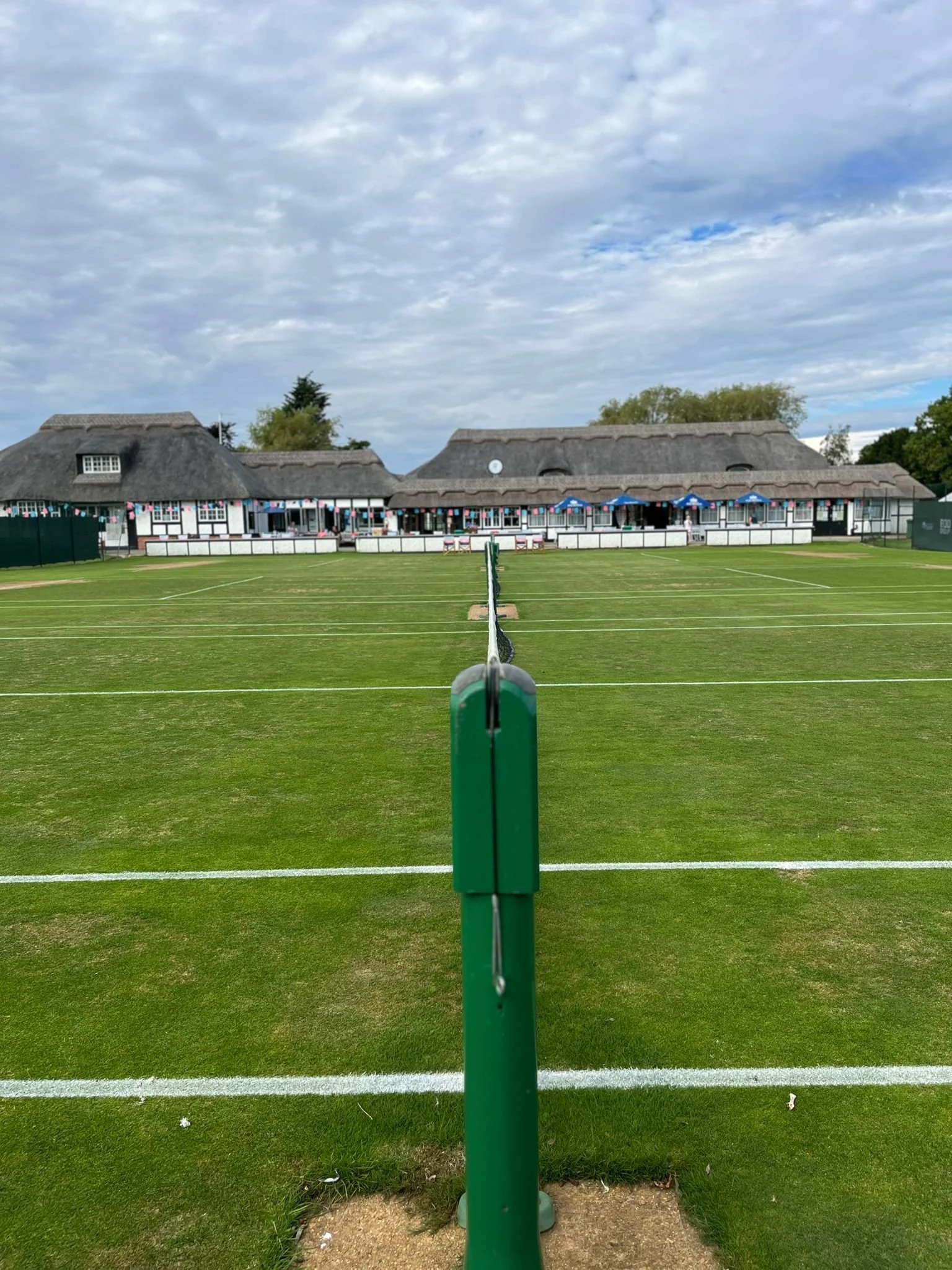 A view of a tennis court with a net in the center, green grass, and a large building with a thatched roof in the background, decorated with flags and umbrellas.