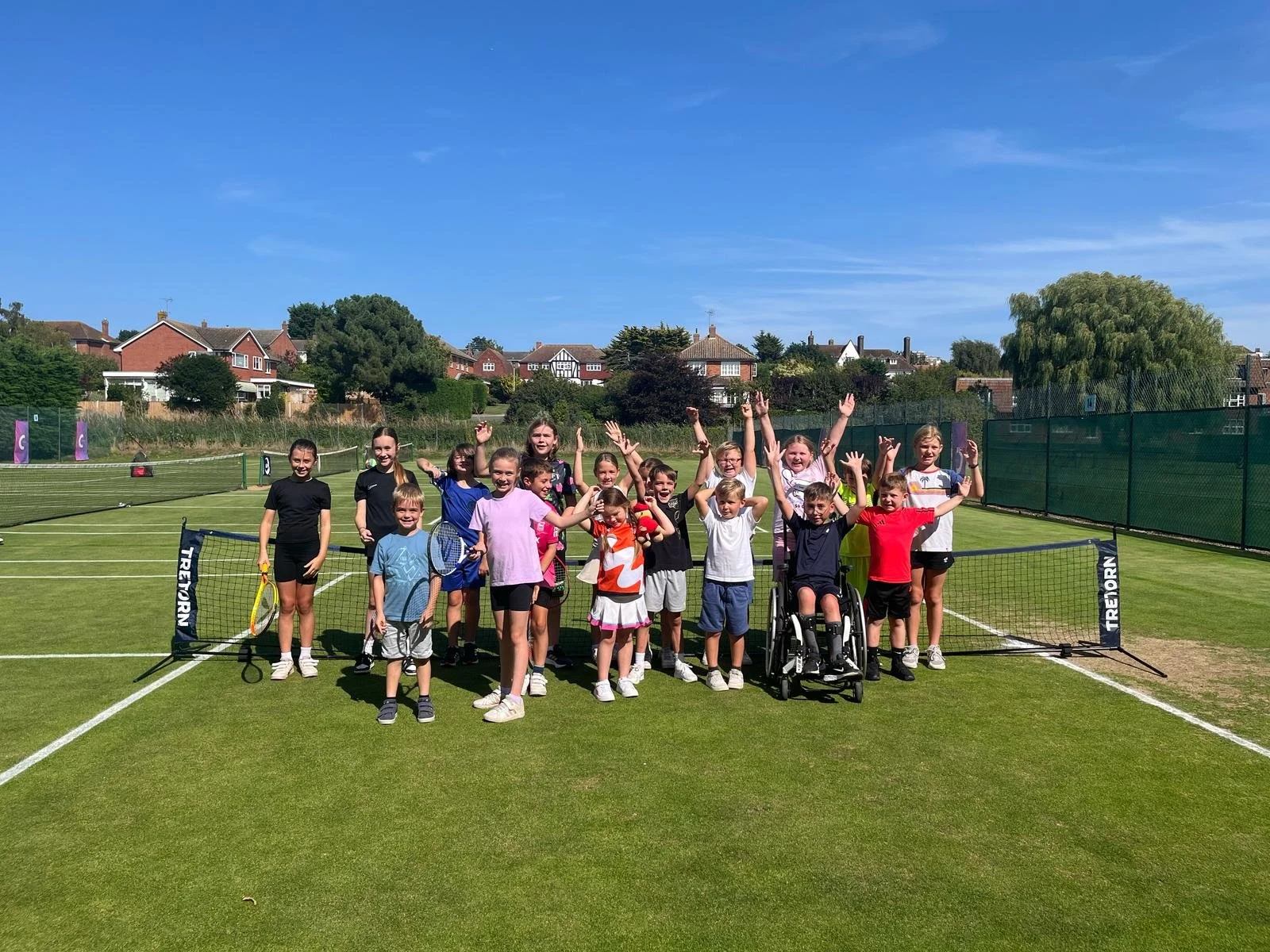 Group of children on a tennis court celebrating after playing tennis, with some raising their arms and smiling under a clear blue sky.