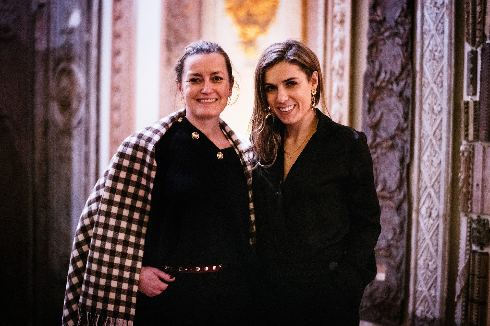 Deux femmes souriantes posent ensemble dans un lieu décoré avec des murs ornés et reliefs, l'une porte une veste à carreaux sur les épaules et l'autre une veste noire.
