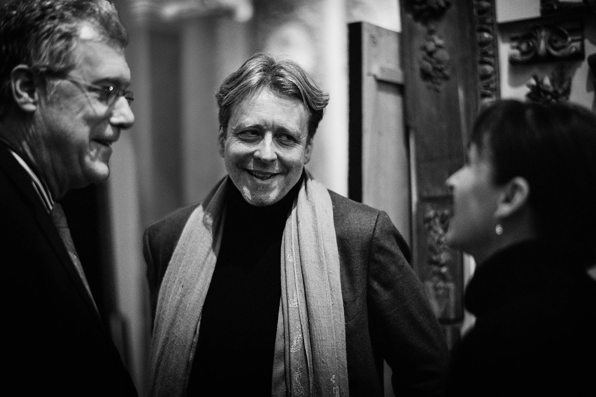 Trois personnes souriantes en conversation, deux hommes et une femme, dans un espace intérieur. L'homme au centre porte une écharpe et sourit, tandis que les deux autres écoutent attentivement.