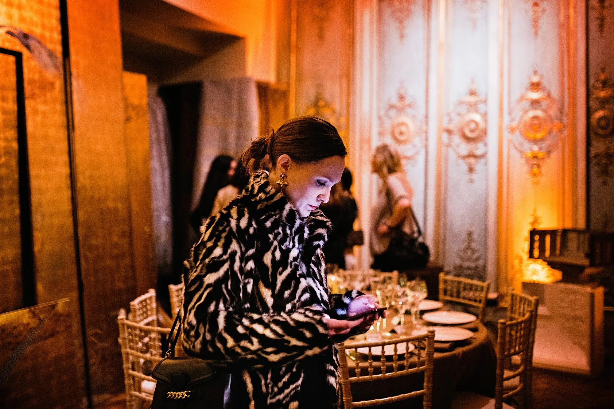 Une femme portant un manteau à motif zèbre regarde son téléphone dans une salle élégante et bien décorée avec des lustres et des meubles en bois doré.