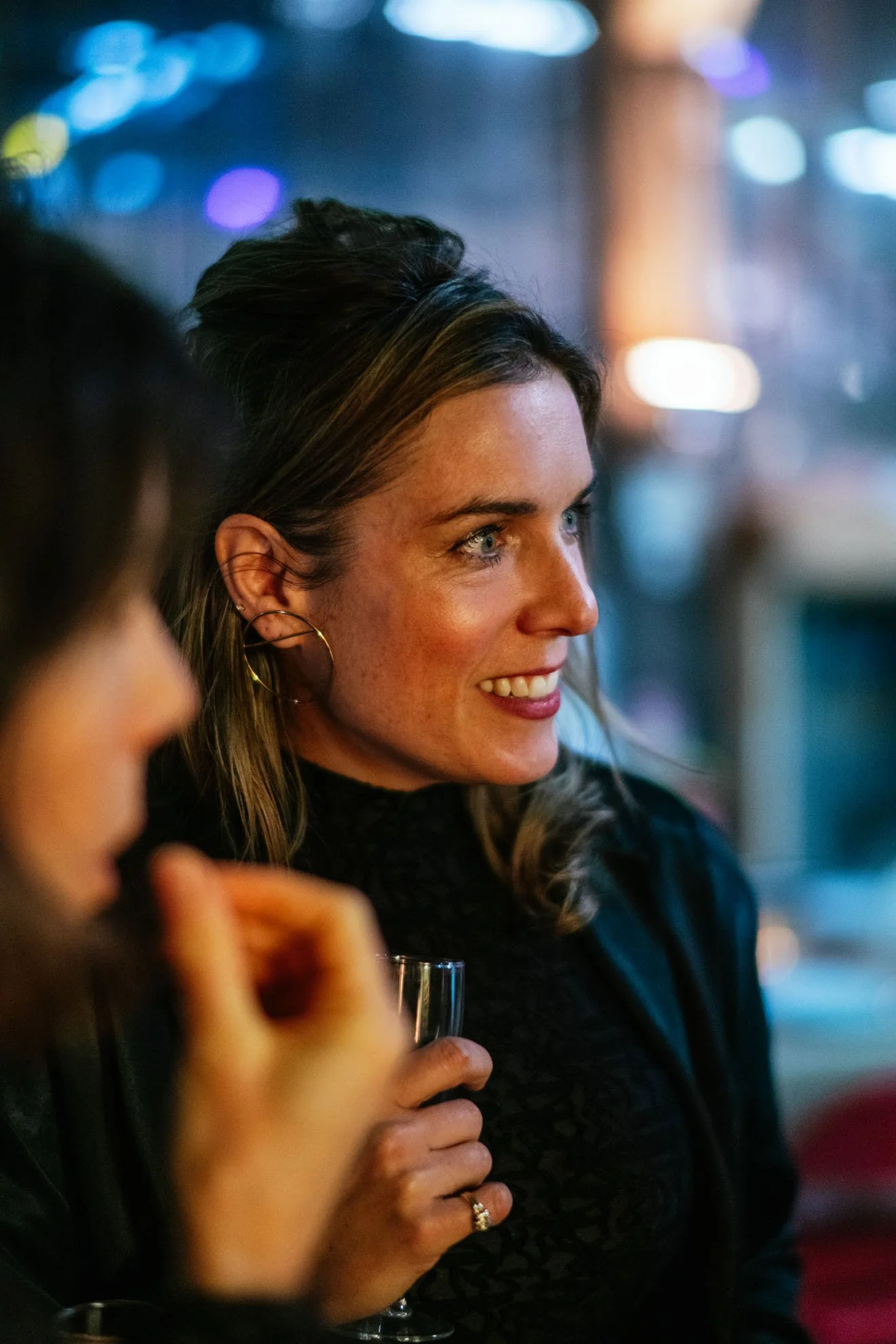 Une femme souriante tenant un verre, dans un environnement éclairé aux couleurs chaudes et bleues.