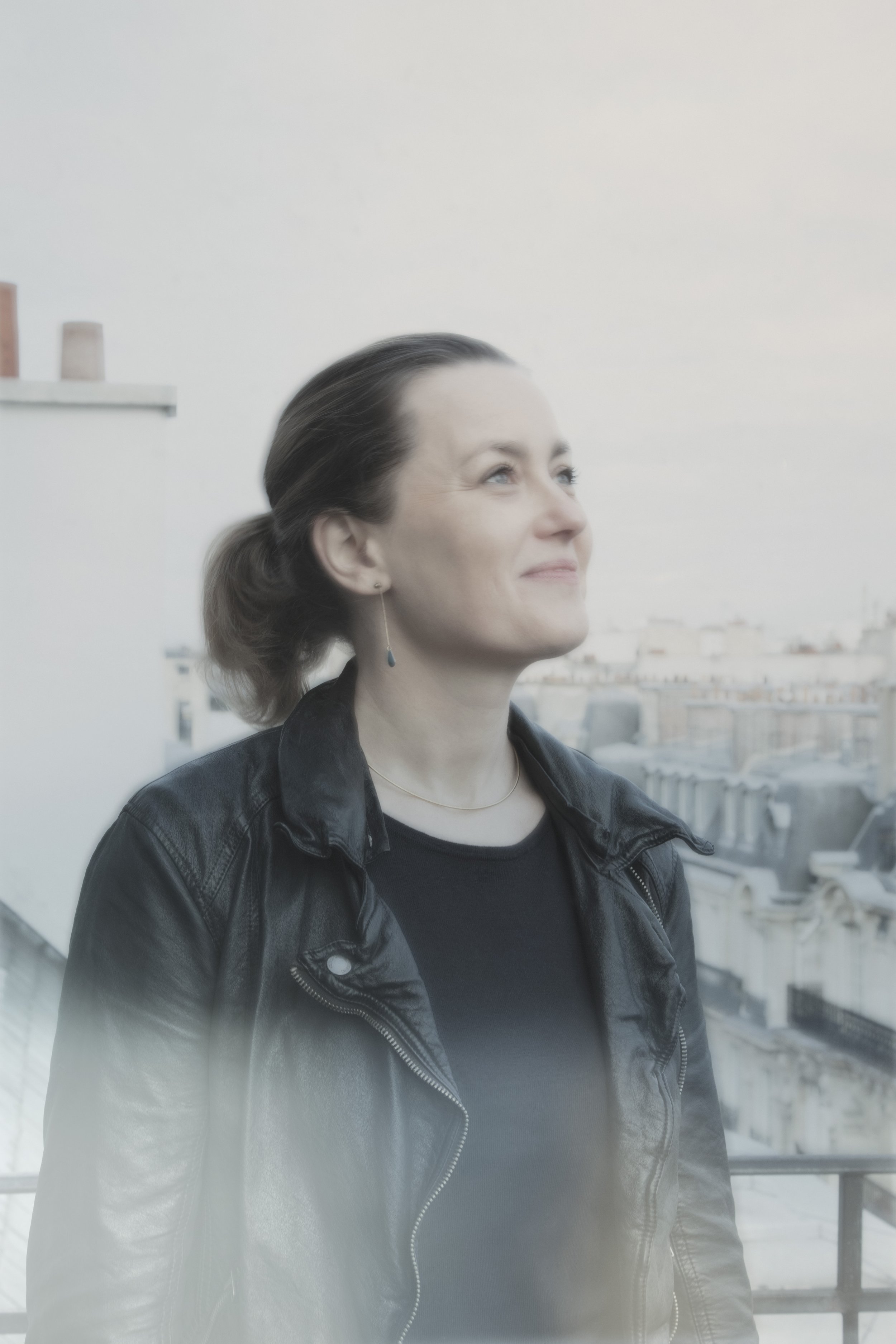 Femme souriante portant un veste en cuir noir, regardant vers le ciel, en arrière-plan un bâtiment parisien avec des toits et des structures urbaines.