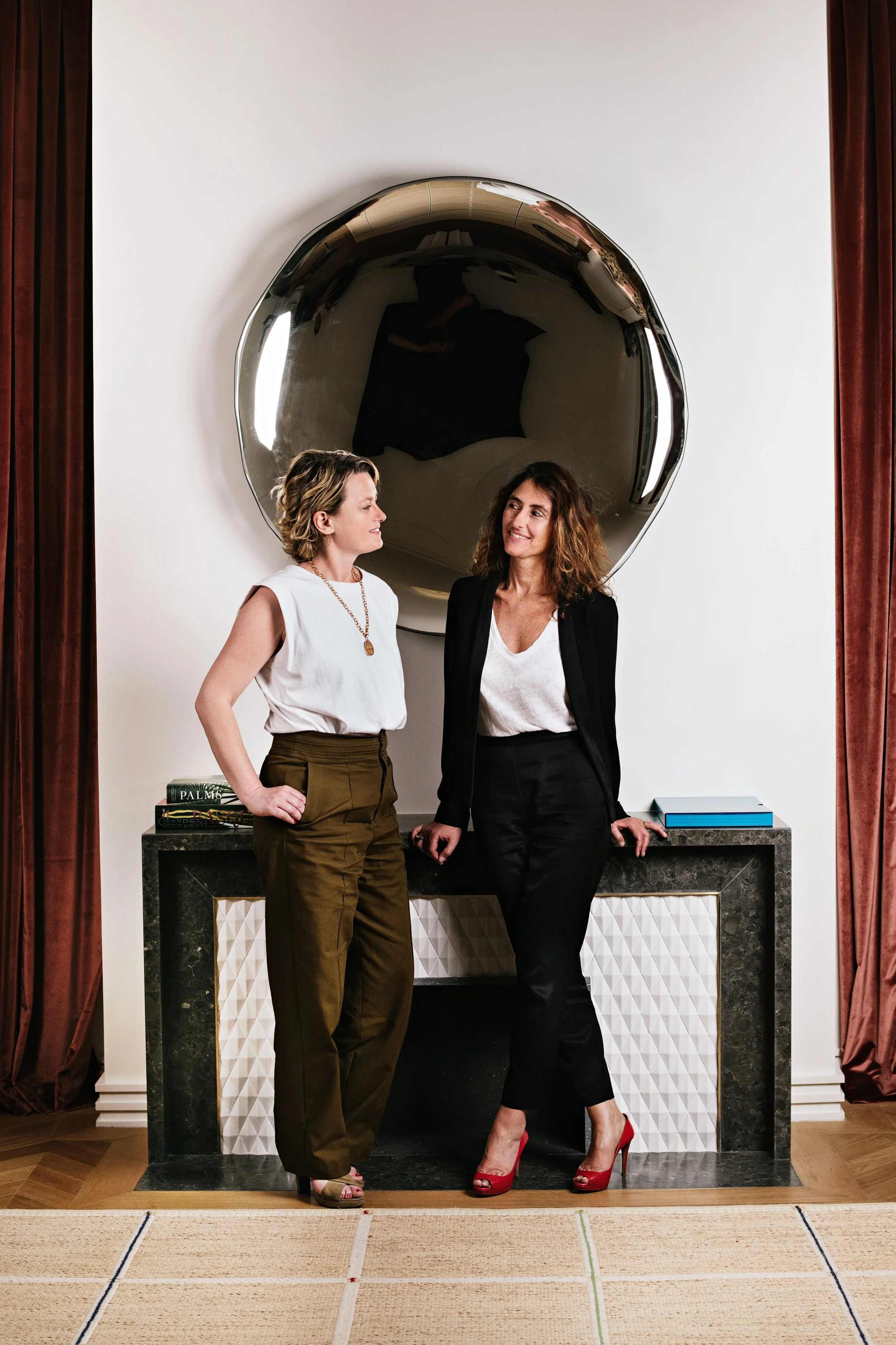 Deux femmes discutant devant une cheminée en marbre sombre avec de la décoration blanche en relief, sous un grand miroir rond en métal, dans une pièce élégante avec des rideaux en velours marron.