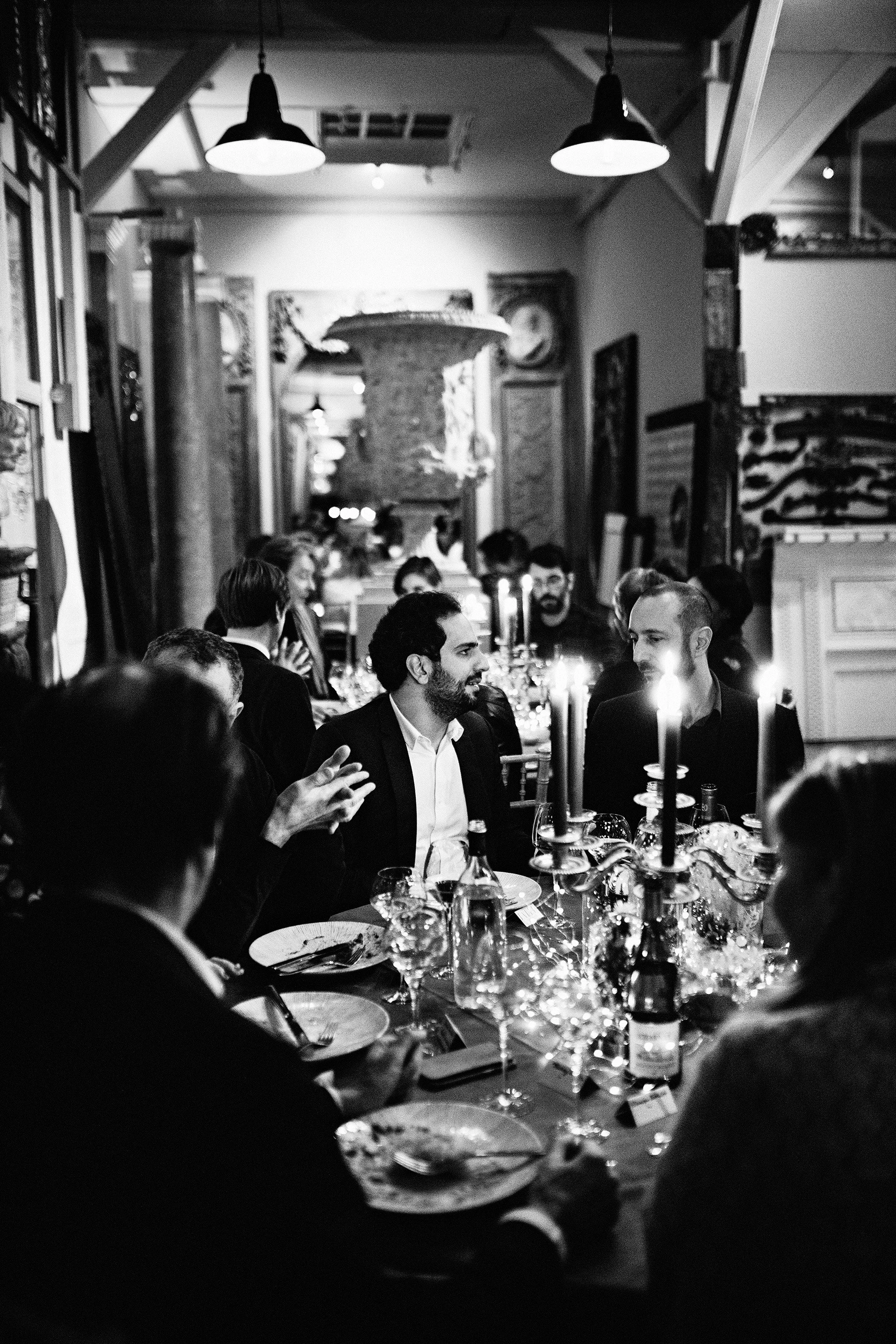 Un groupe de personnes assises autour d'une longue table lors d'un dîner formel dans un restaurant ou une salle de banquet, décorée avec des bougies et des décorations festives, en noir et blanc.