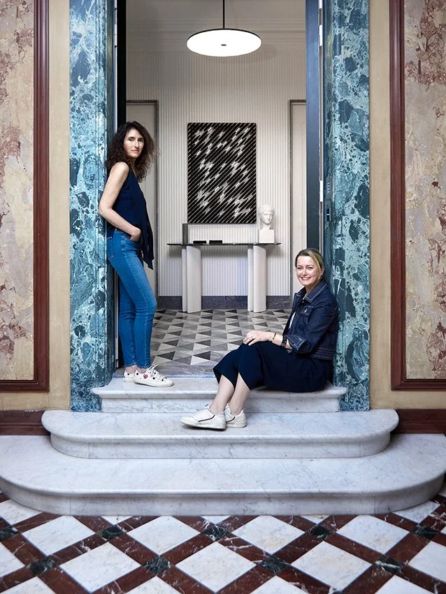 Deux jeunes femmes dans une entrée d'immeuble, une debout dans la porte et l'autre assise sur le seuil, avec un décor intérieur moderne et artistique en arrière-plan.