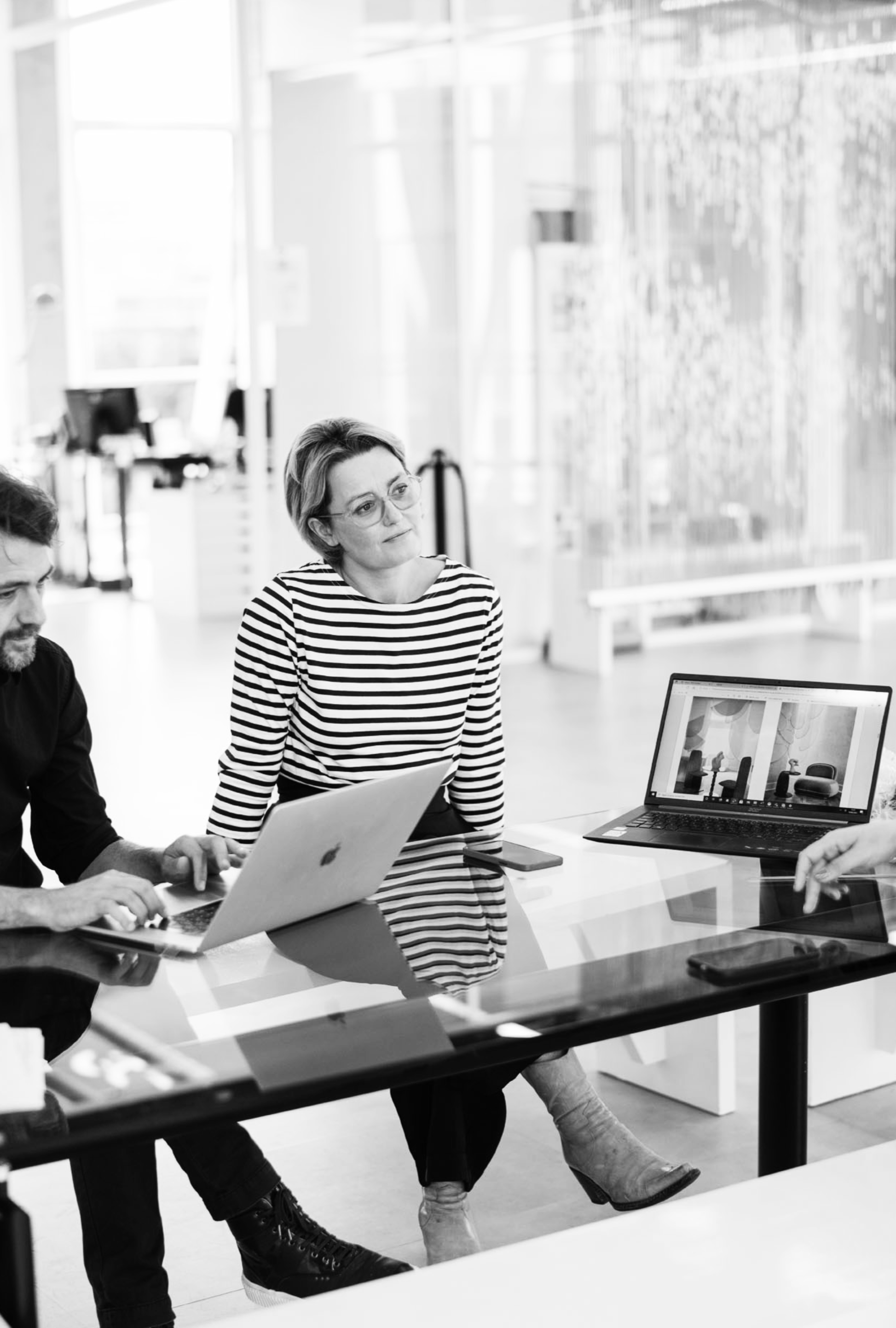 Trois personnes en réunion dans un espace de travail moderne, deux avec des ordinateurs portables ouverts, une femme en chemise à rayures, assise sur un bureau en verre, regardant attentivement.