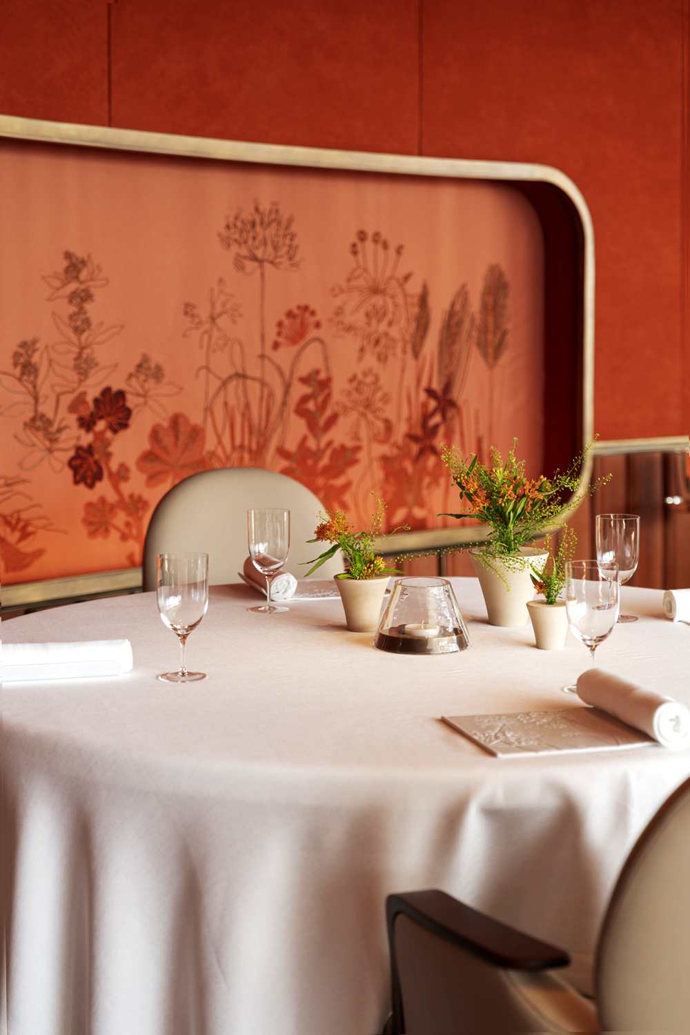 Table de banquet élégante avec nappe blanche, décor floral avec deux petits pots de fleurs, un centre de table avec bougie, verres à vin, assiettes et serviettes, dans une salle avec mur orange à motif floral.