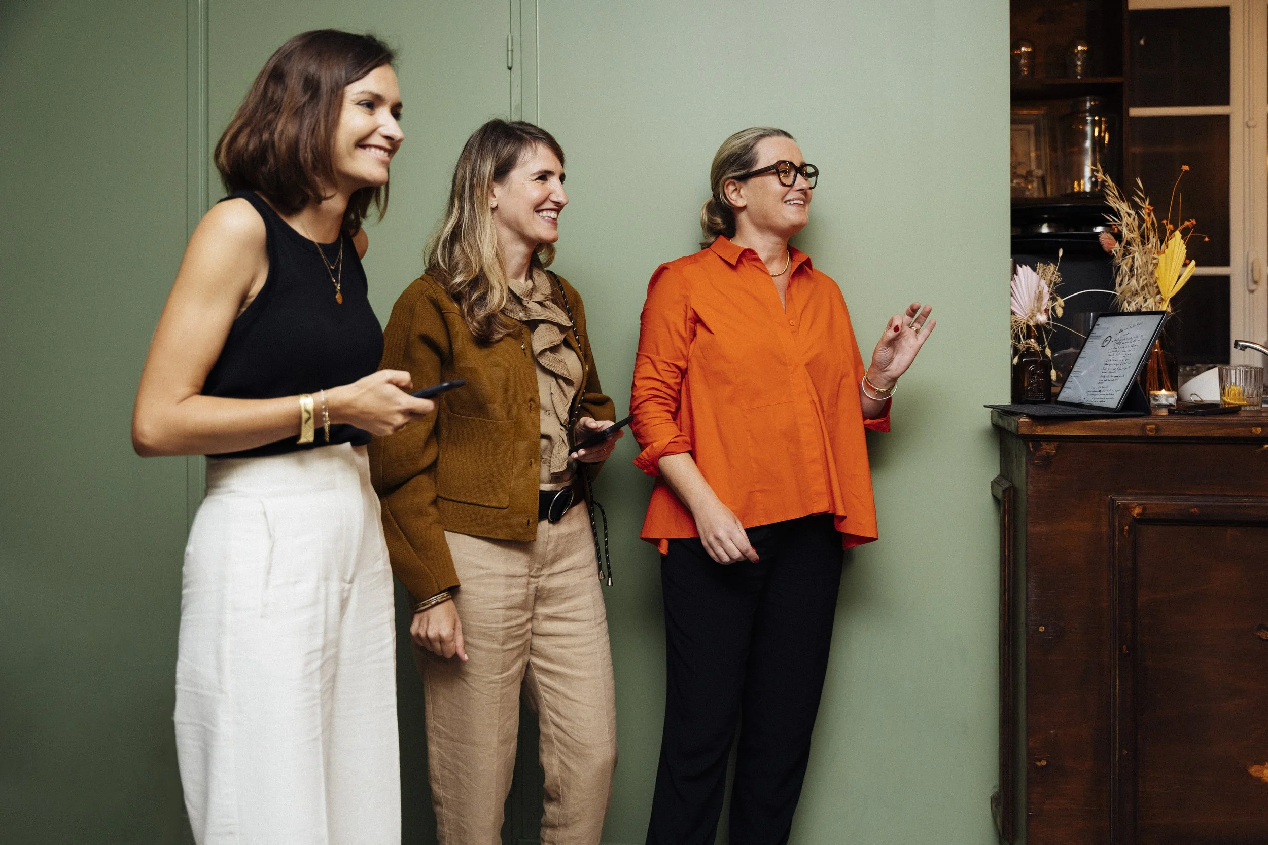 Trois femmes debout près d'un mur vert, regardant une tablette posée sur un meuble en bois, en train de discuter et de sourire.