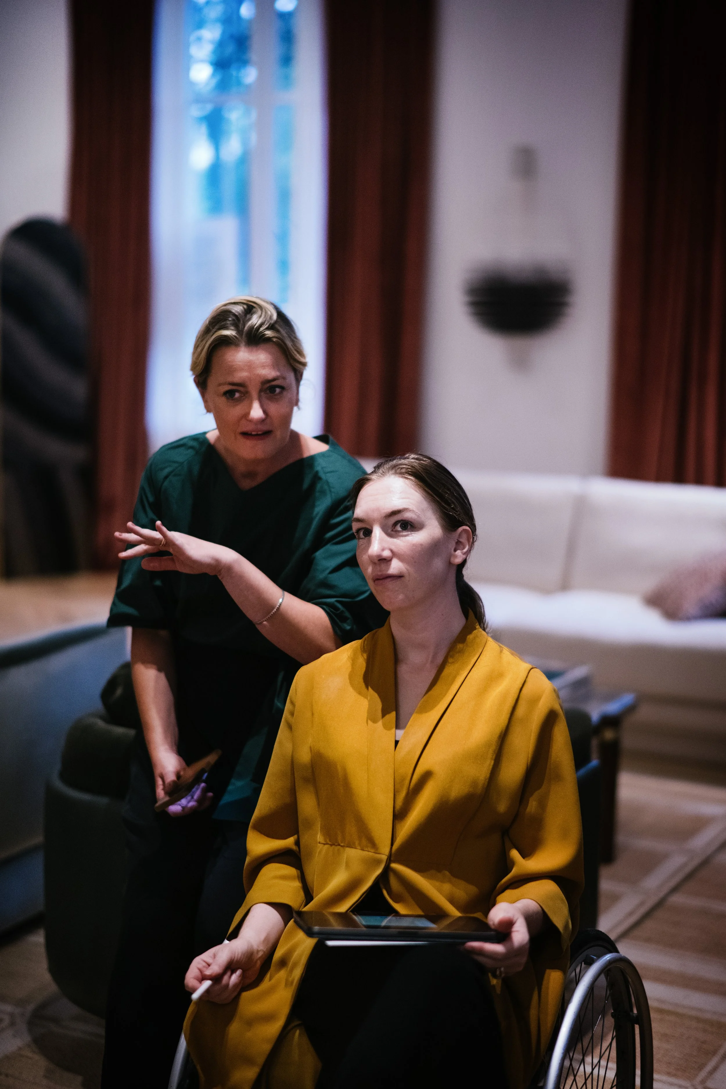 Deux femmes, dont une en fauteuil roulant, semblent discuter ou écouter quelqu'un dans un salon. La femme en fauteuil porte un manteau jaune, et l'autre une tenue verte.