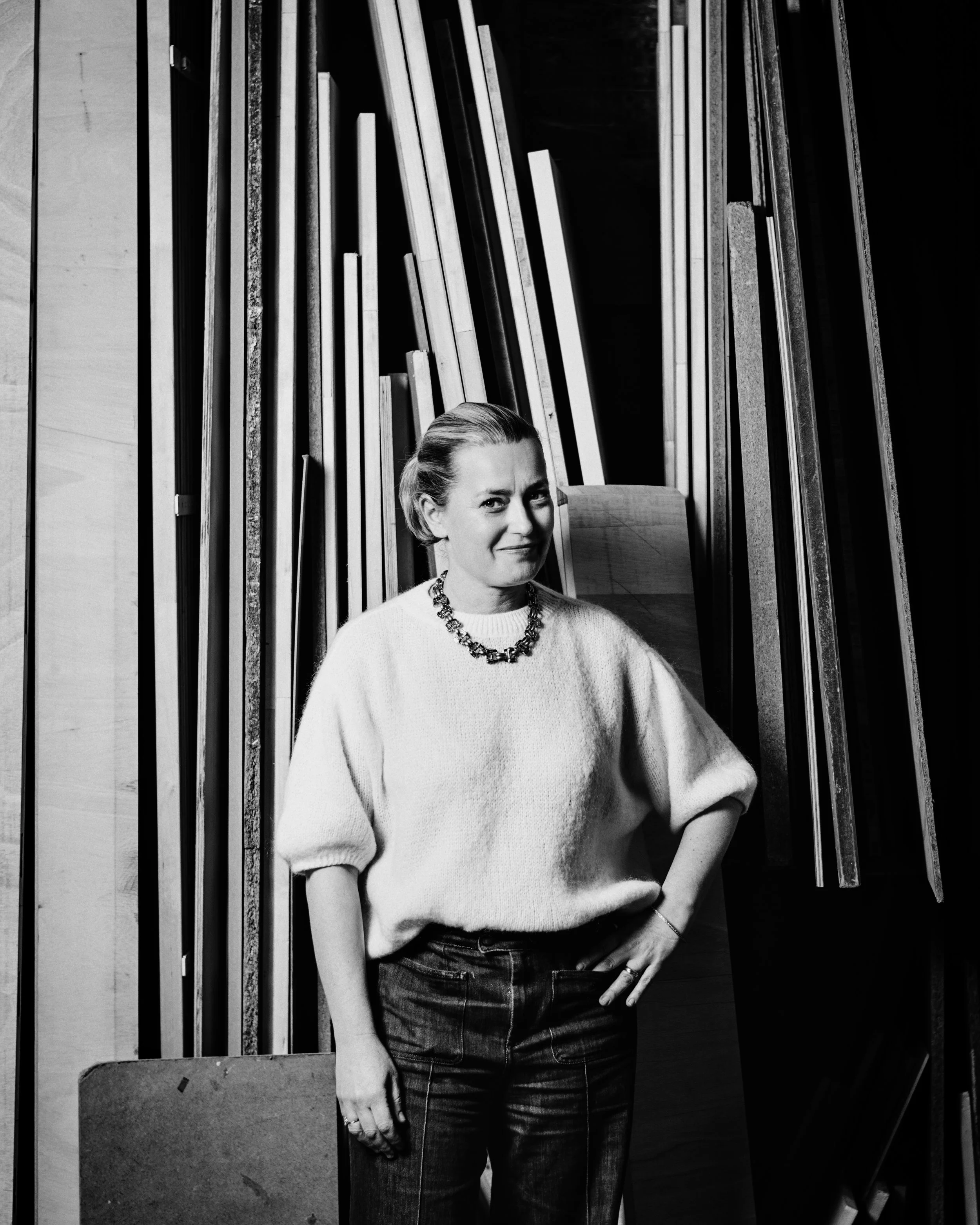 Une femme posant dans un atelier, en noir et blanc, avec des planches de bois visibles en arrière-plan.