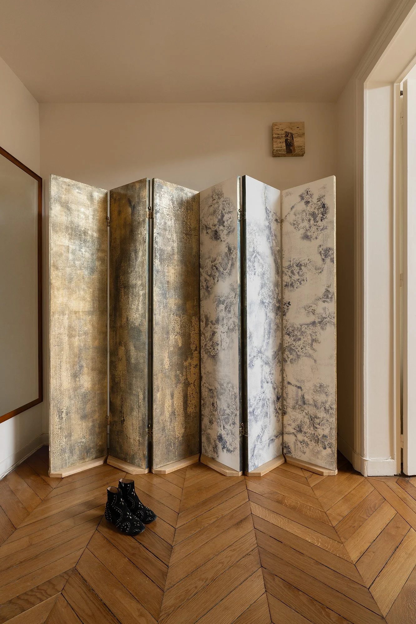 Un paravent décoratif à quatre volets avec des motifs marbrés en or et en blanc, placé dans un intérieur avec un plancher en bois et une paire de bottines noires devant.