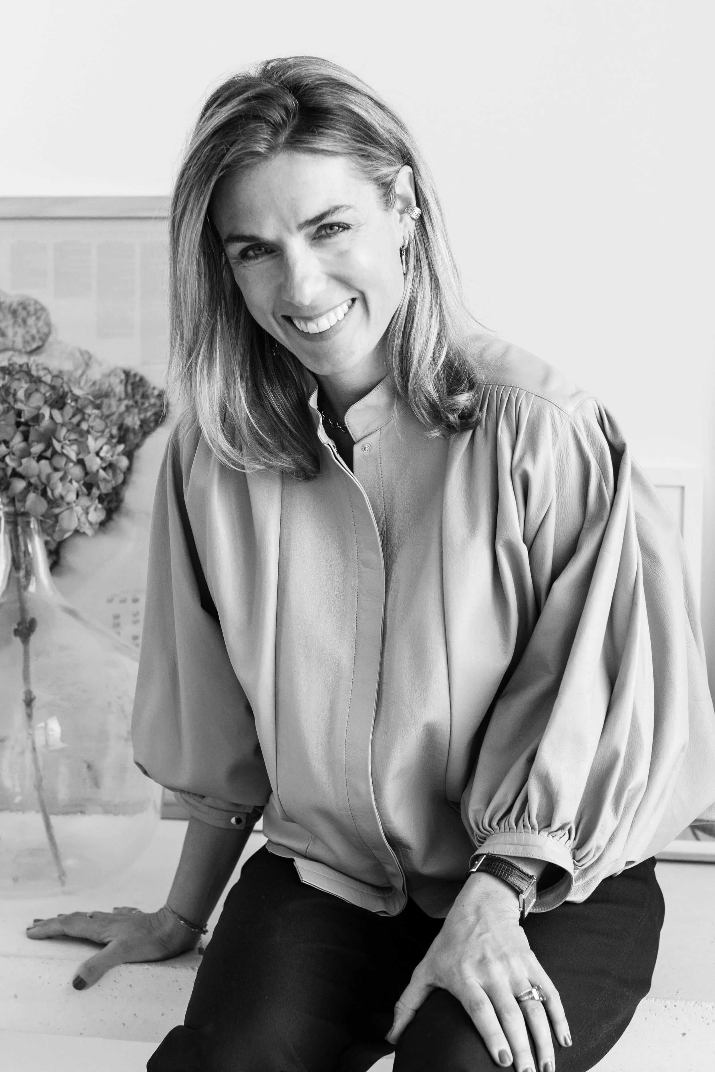 Une femme souriante avec des cheveux mi-longs, portant une blouse à manches bouffantes et un bracelet, assise sur un comptoir, dans un environnement intérieur.