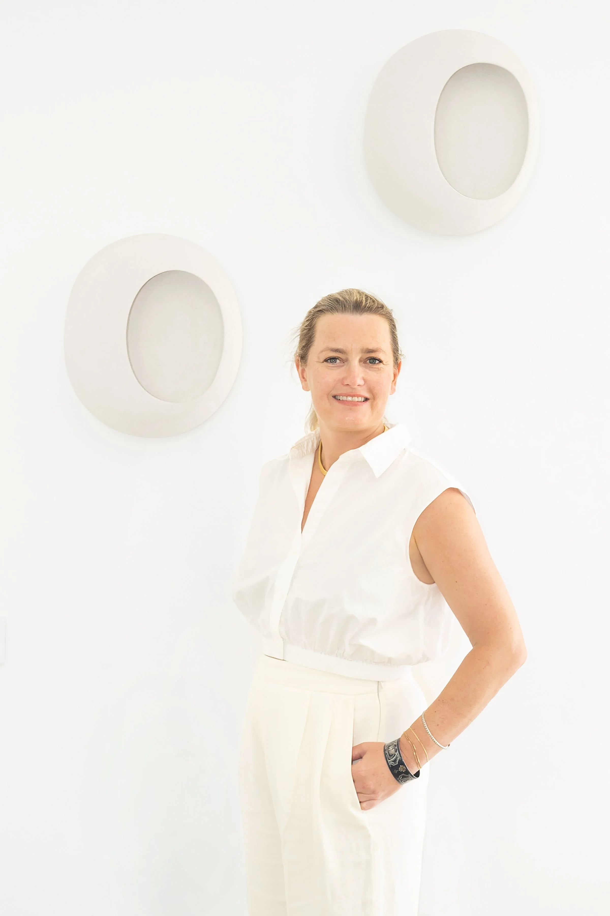 Femme souriante en tenue blanche, debout devant un mur blanc avec deux décorations circulaires.