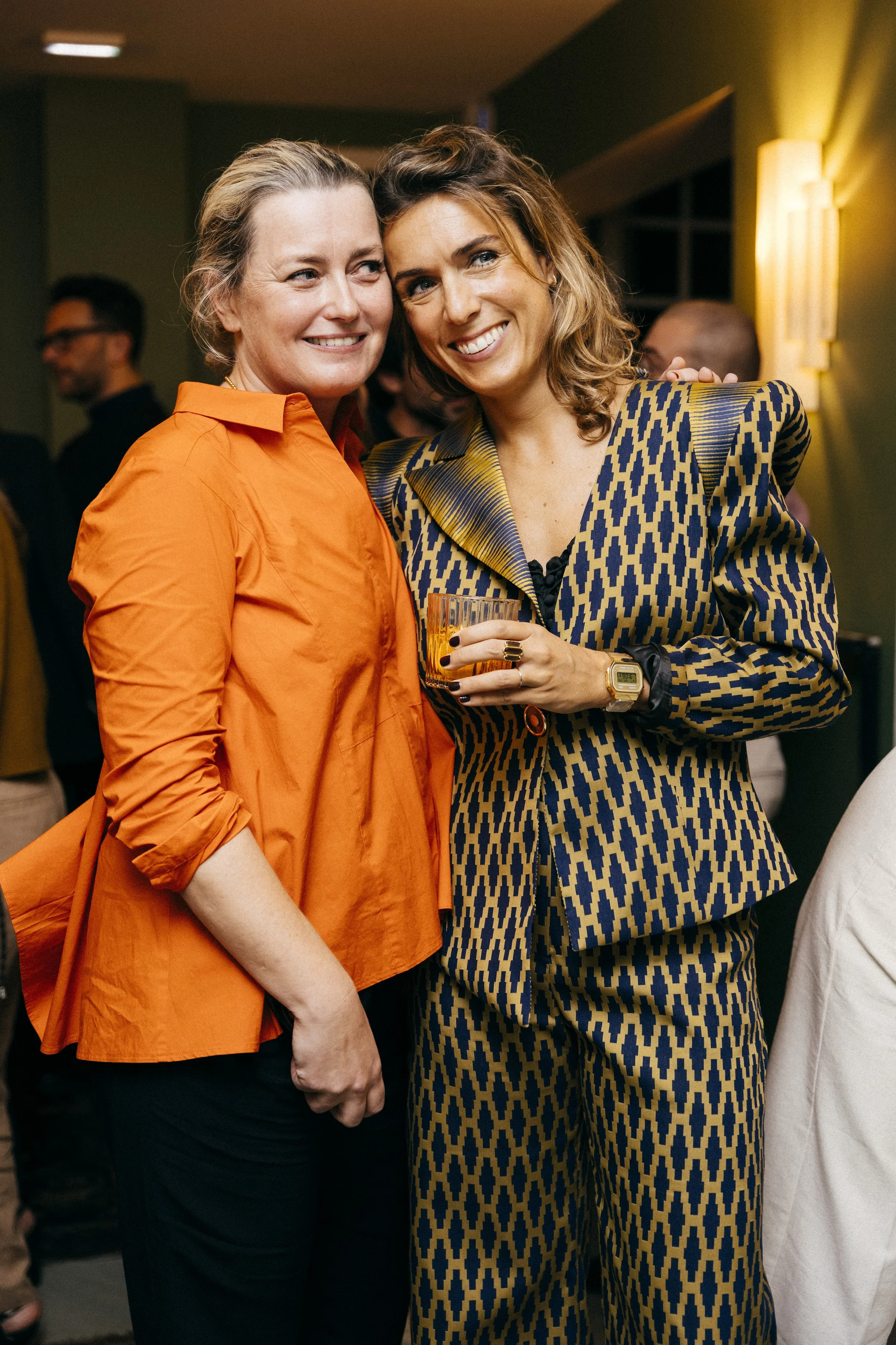 Deux femmes souriantes posent lors d'une fête, l'une porte une chemise orange, l'autre un costume à motif bleu et jaune, tenant un verre.