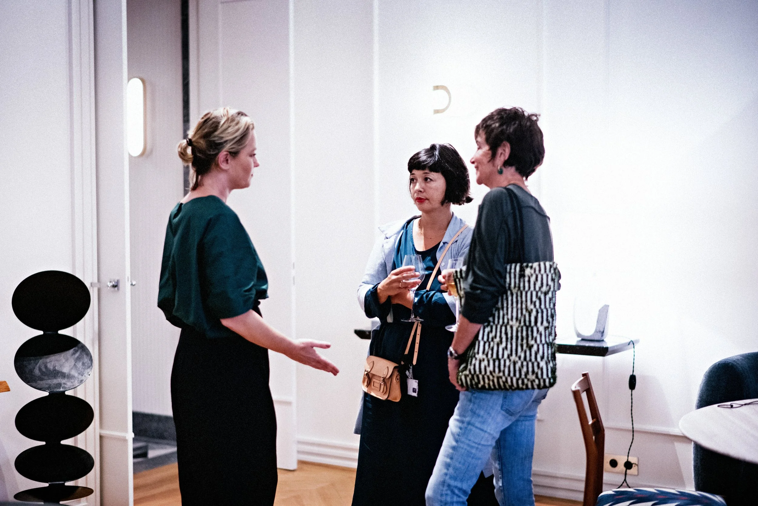 Trois femmes discutent dans une pièce sobrement décorée, une d'elles parle avec une expression sérieuse, les deux autres écoutent attentivement, tenant des verres à la main.