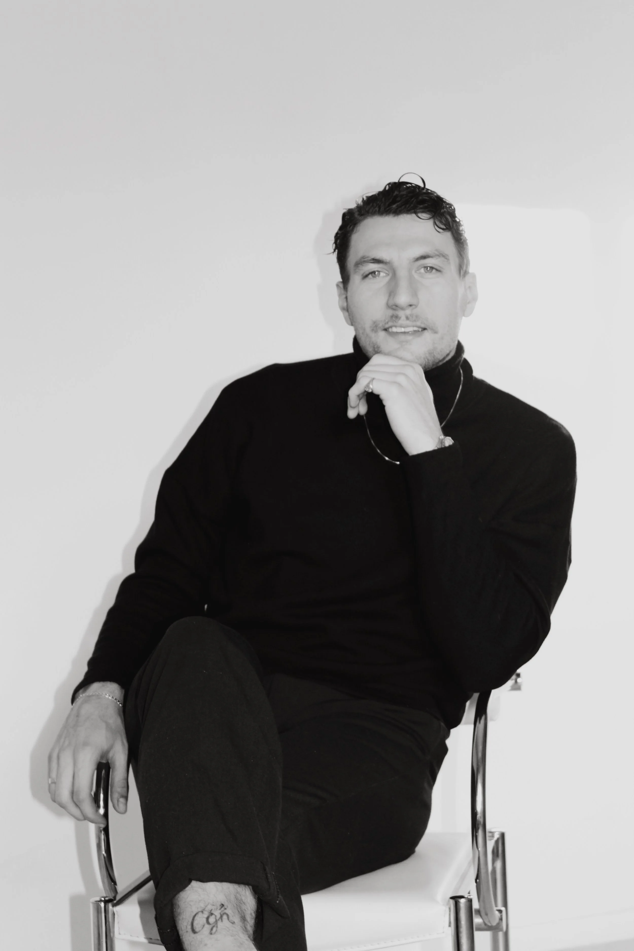 Black and white photograph of a man sitting on a chair against a plain background. He is wearing a black turtleneck sweater and has short, curly hair. He is resting his chin on his right hand, which is near his face, and holding a smartphone in his left hand. His right ankle is resting on his left knee, and he has a tattoo on his left ankle.