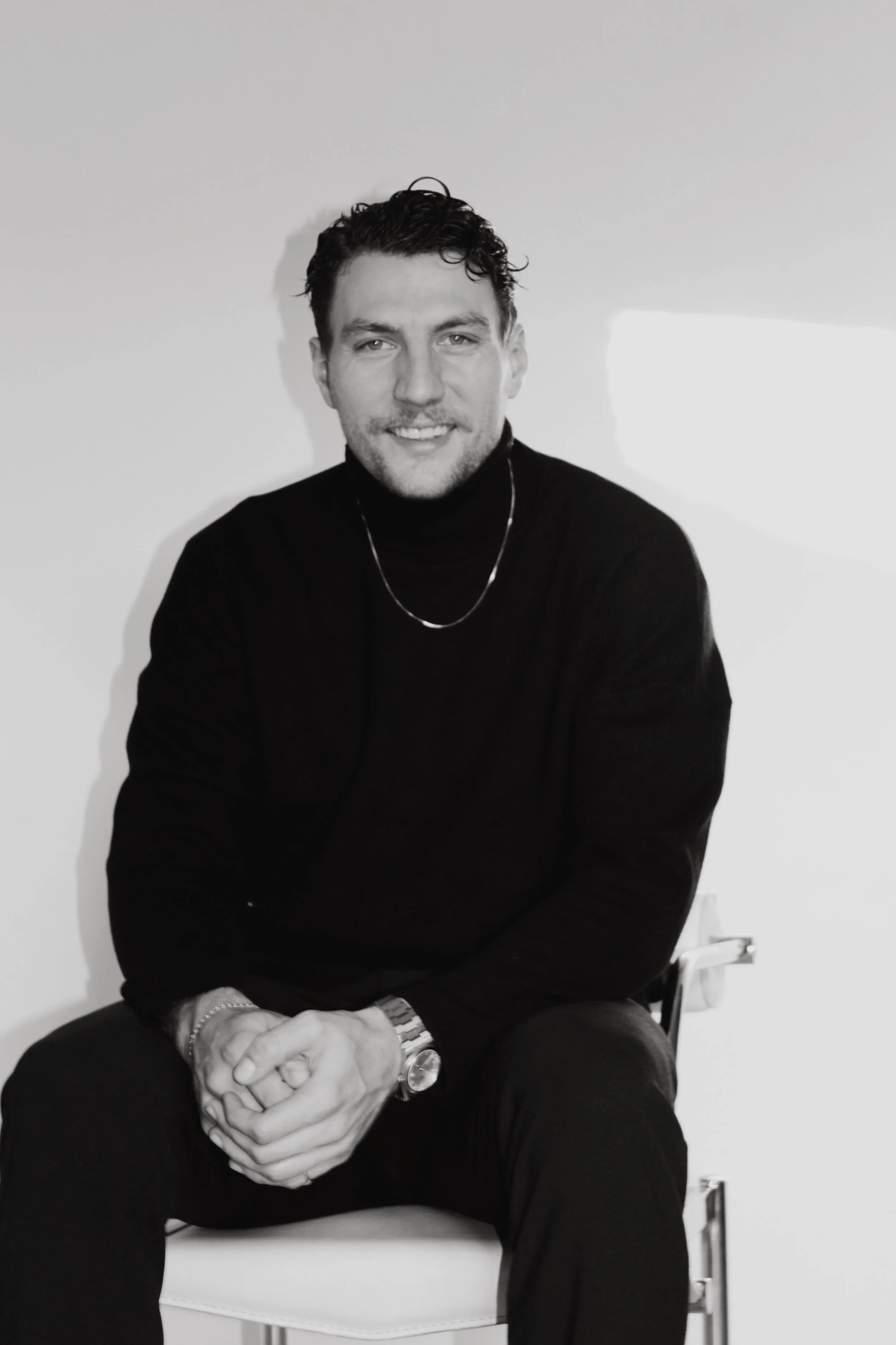 Black and white portrait of a smiling man with short curly hair, wearing a black turtleneck sweater, a chain necklace, a watch, and a bracelet, sitting on a chair against a plain wall.