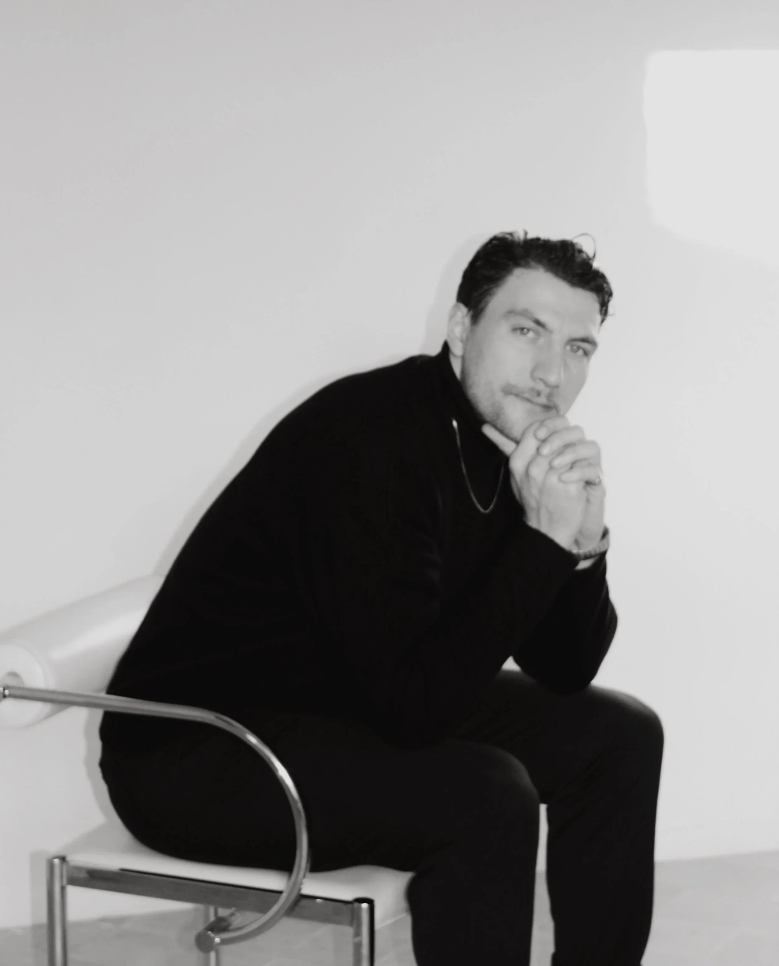 A man with dark hair and beard, dressed in black, sitting on a modern chair with a metal armrest, against a plain white wall, with his hands clasped and resting on his chin, looking at the camera.