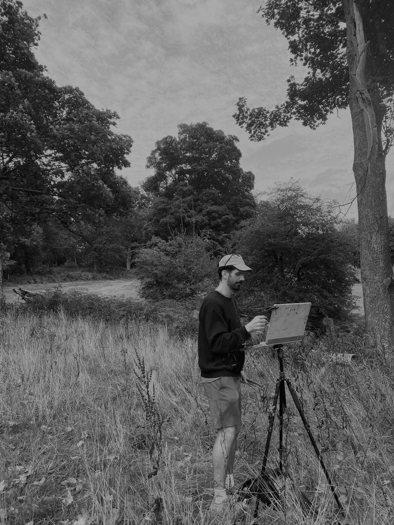 A man standing on a grassy field with trees, using an easel outdoors.