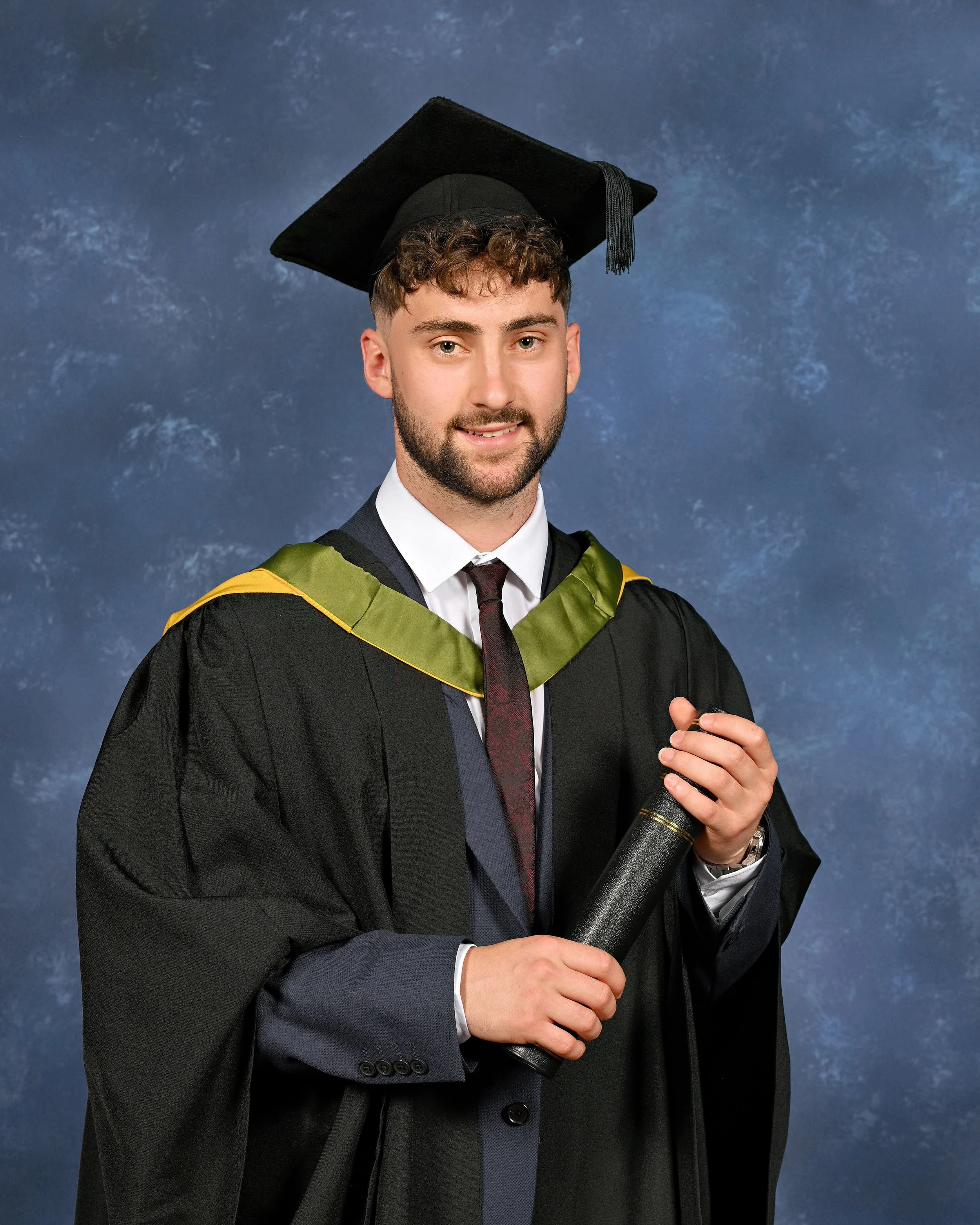 Young man in graduation cap and gown holding diploma, standing against a blue backdrop.