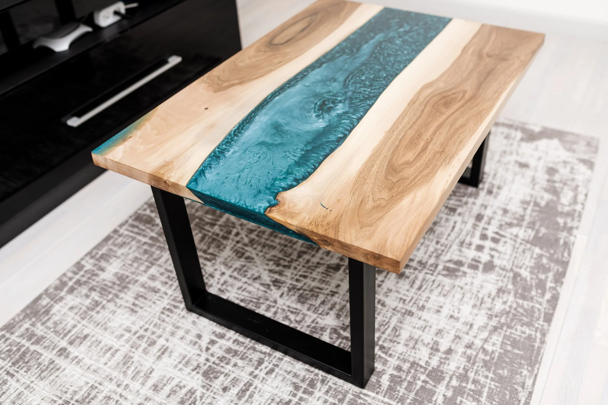 Wooden table with a river-like blue resin inlay on top, black metal legs, placed on a patterned rug.