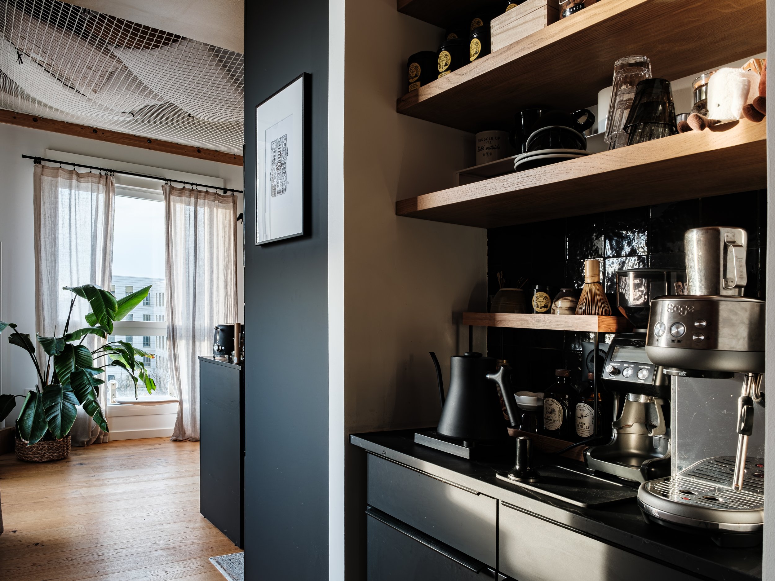 Intérieur d'une cuisine moderne Schmidt avec un corner coffee shop avec étagères en chêne massif, une machine à espresso Sage, et des tasses. Une grande fenêtre avec des rideaux en tissu léger, un grand plante verte près de la fenêtre 