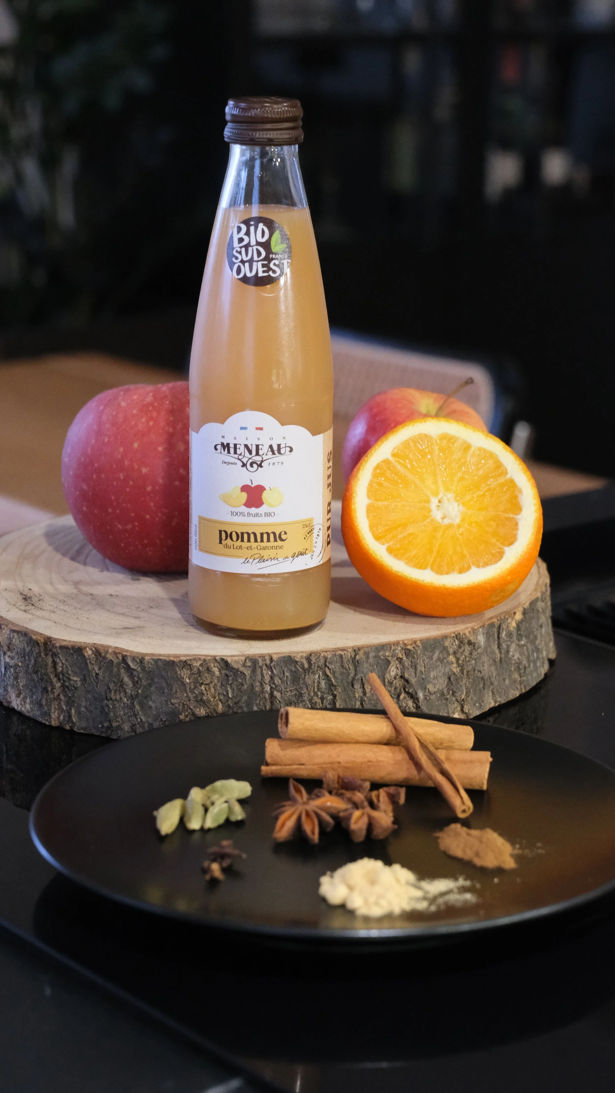 Une bouteille de jus de pomme bio, une pomme rouge, une orange coupée en deux, un rondin de bois, et des épices dont de la cardamome, de la cannelle, de la muscade, du gingembre en poudre, des clous de girofle et du poivre noir, sur une planche en bo