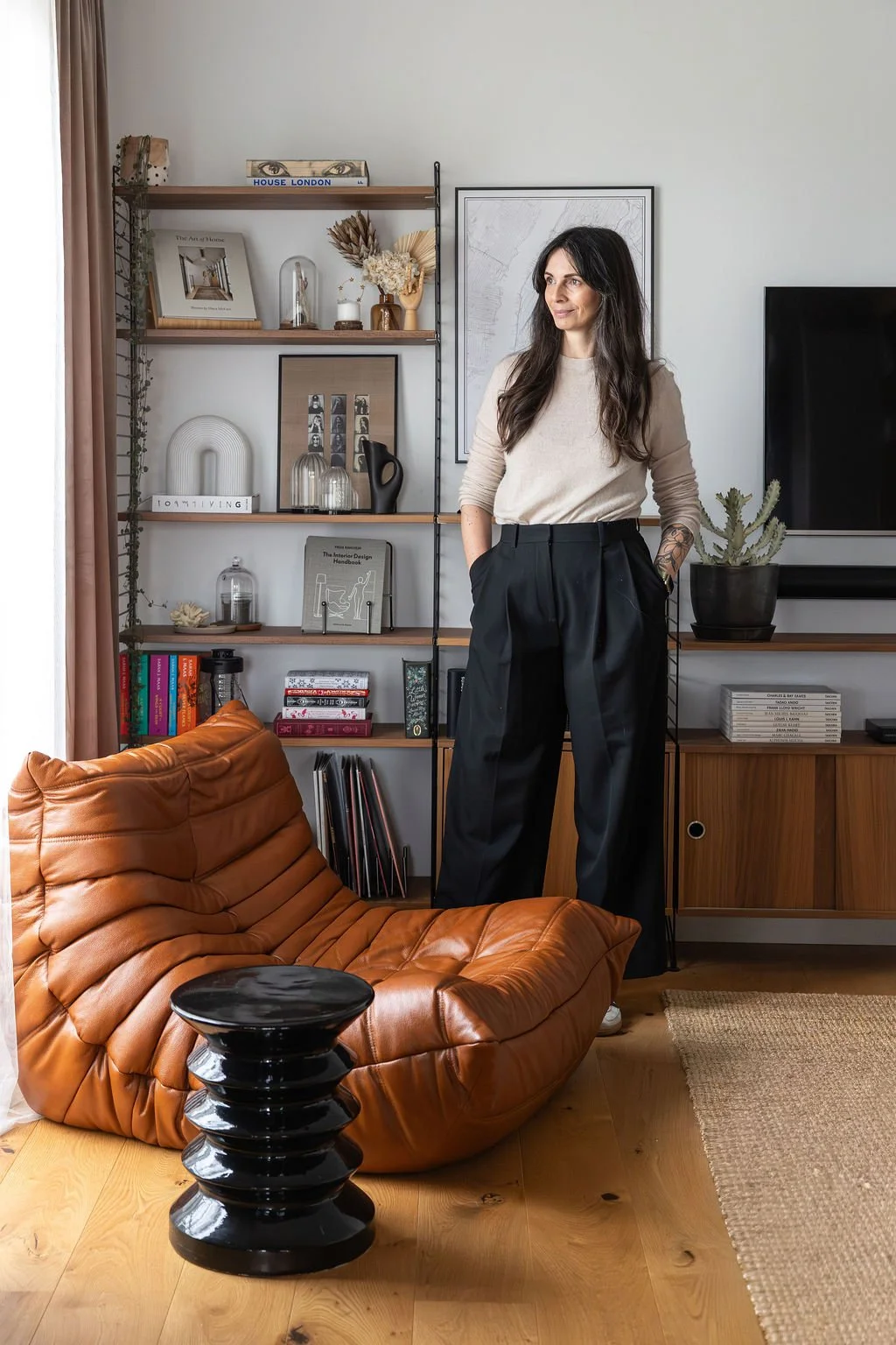 Une femme décoratrice d'intérieur à Bordeaux debout dans un salon moderne avec une composition String Furniture en noyer, une étagère avec des décorations et un fauteuil en cuir marron modèle Togo de ligne roset.