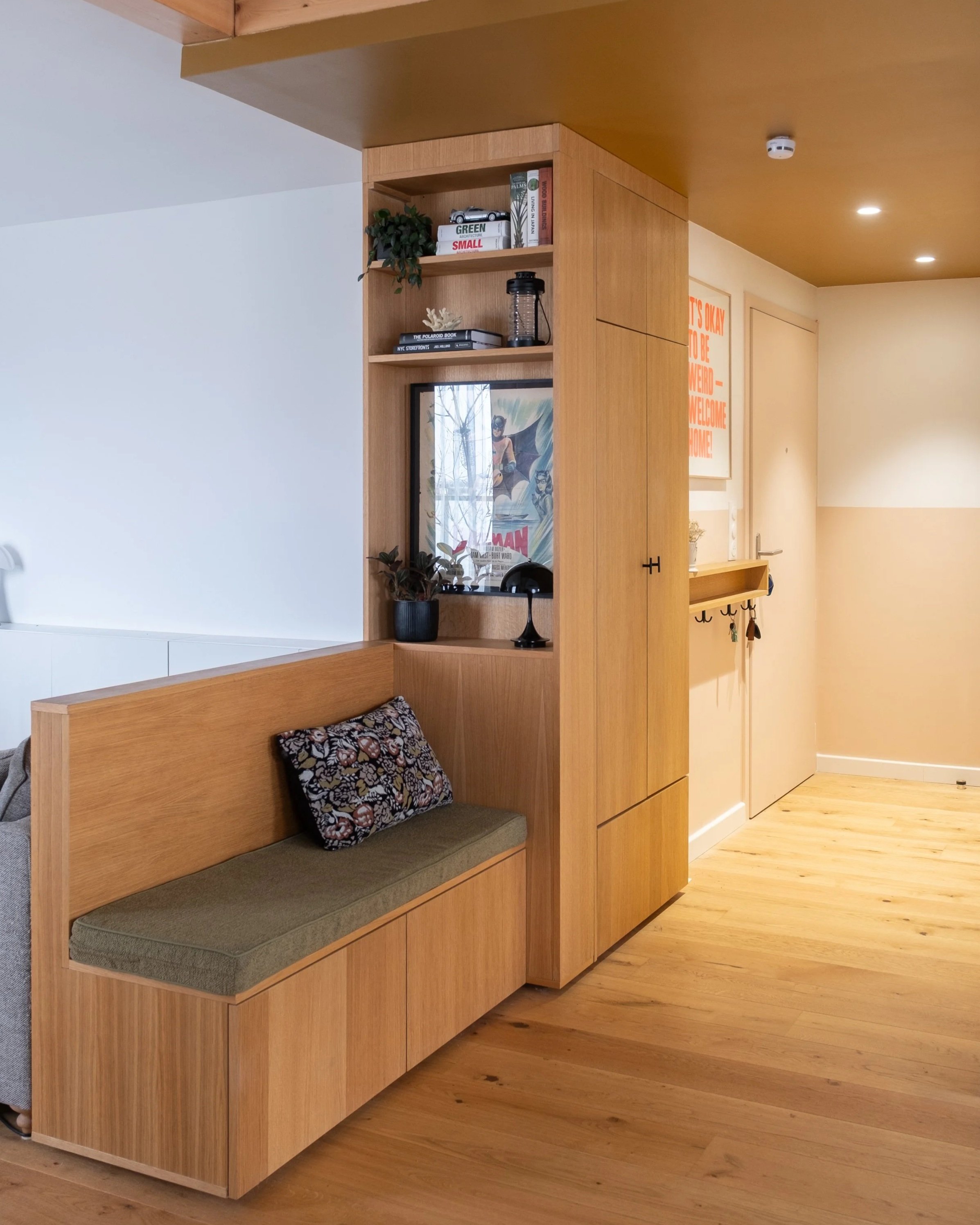 Un coin d'entrée dans un appartement avec un meuble en bois clair comportant une banquette avec un coussin, une petite étagère avec des objets décoratifs, des livres, une plante et une lampe. À l'arrière, un mur avec une affiche orange et blanche, et une porte fermée.