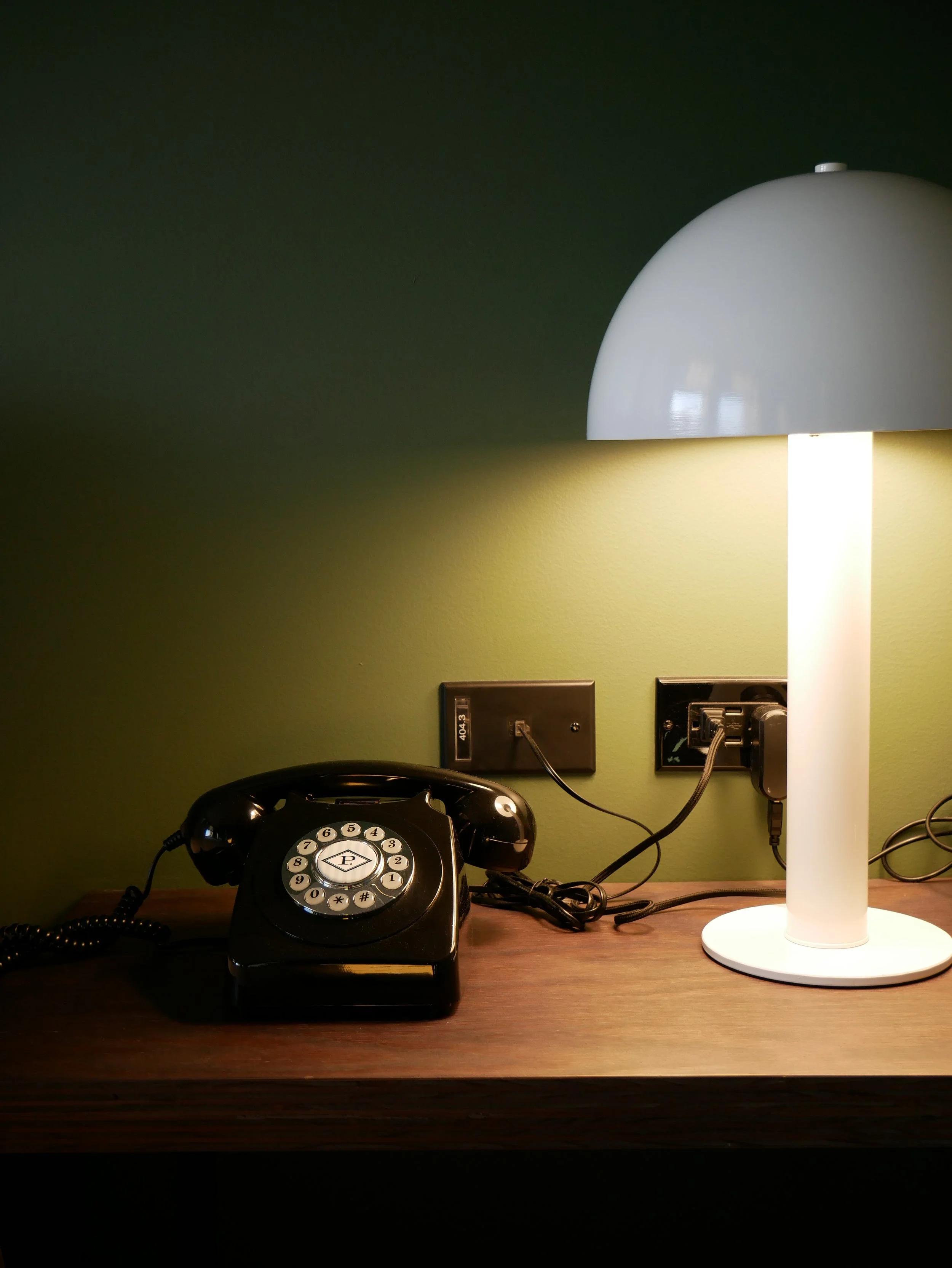 Une table en bois avec une lampe blanche à droite, une prise électrique et un téléphone rétro noir à gauche.