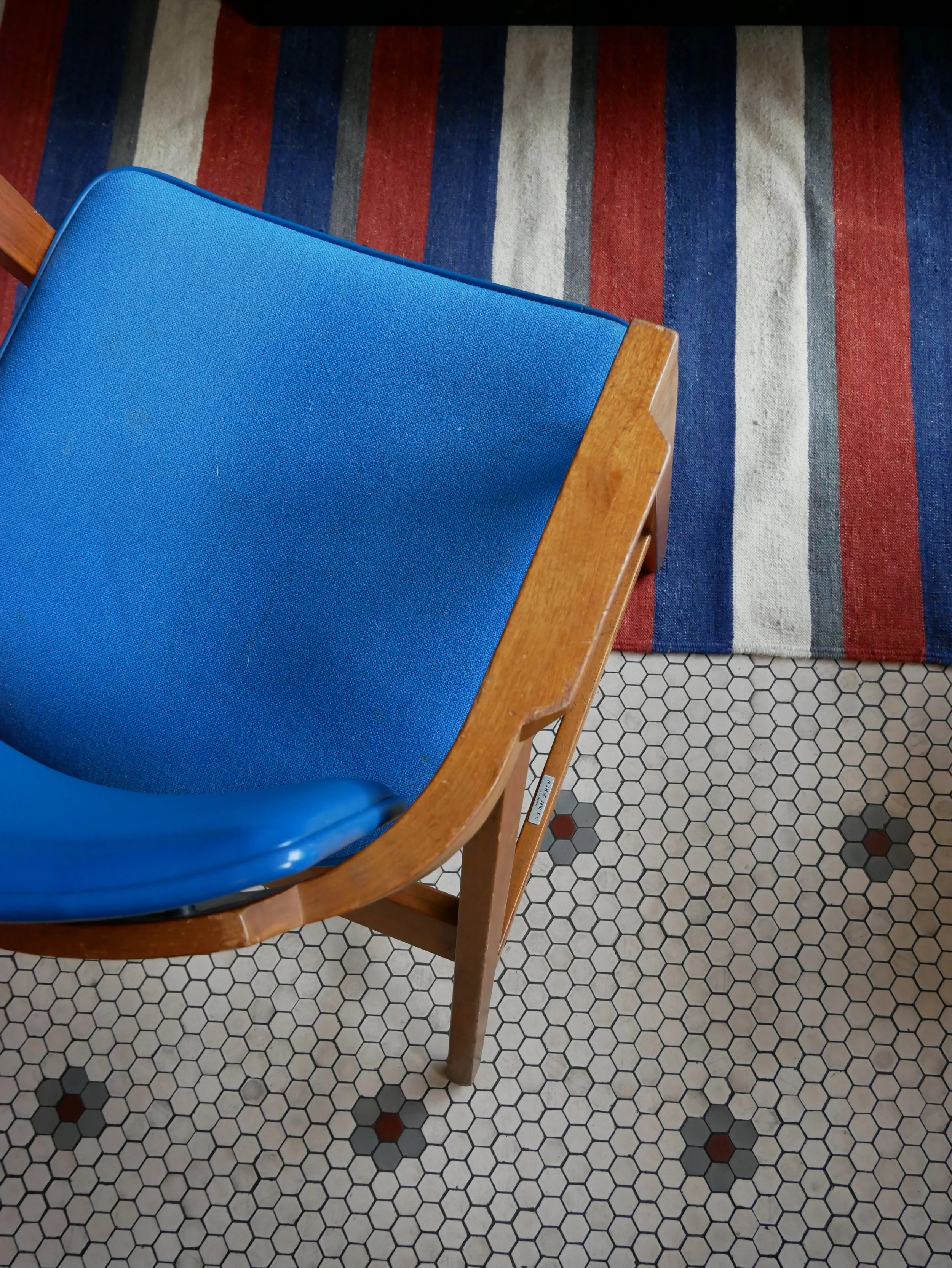 Chaise en bois avec assise et dossier bleus, sur un sol en carrelage hexagonal blanc avec quelques carrés noirs et roux, et un tapis rayé en arrière-plan avec des bandes rouges, grises, beiges et bleues.