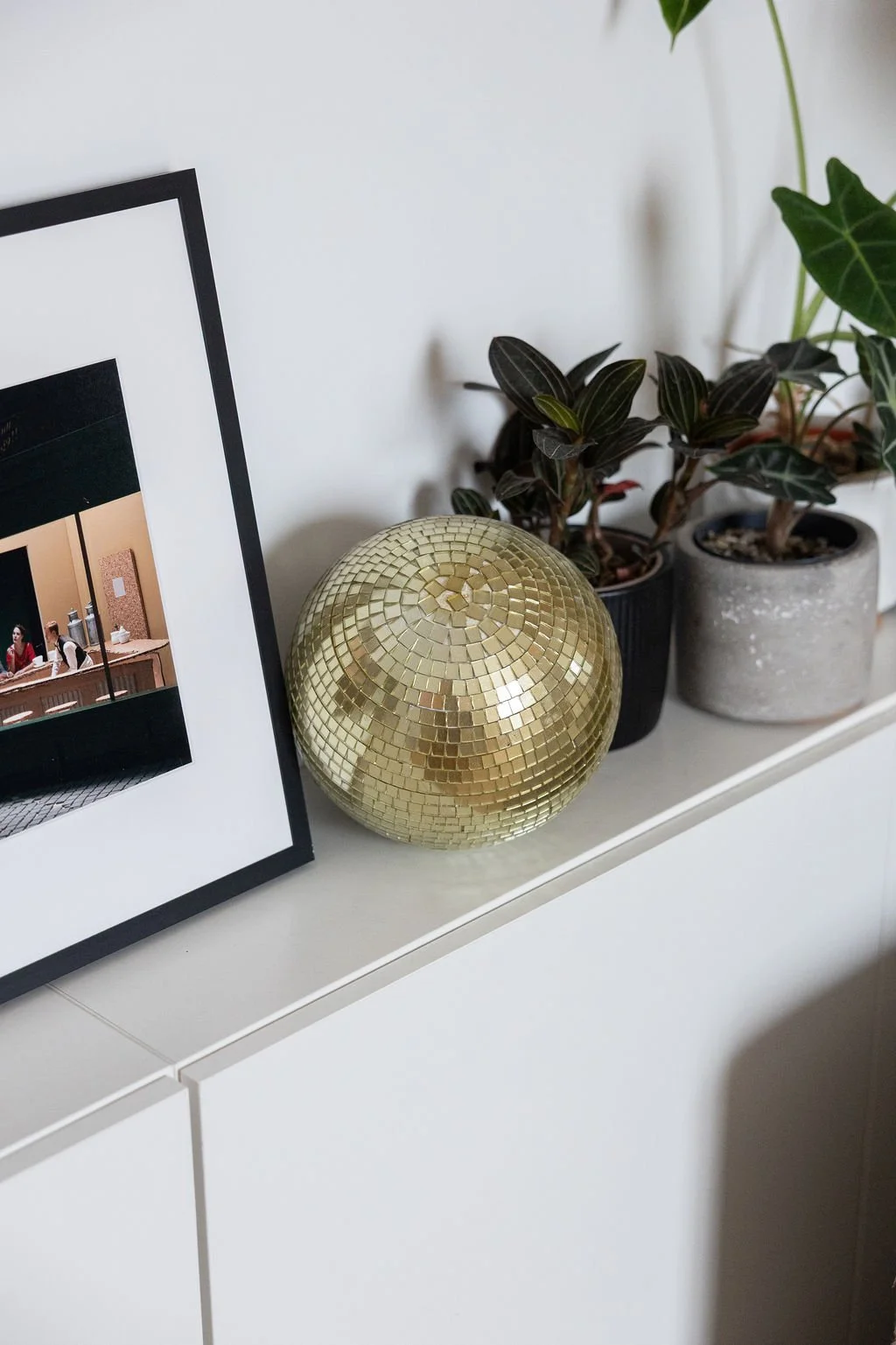 Une console blanche avec un cadre photo noir, une boule à facette dorée, et deux pots de plantes vertes.