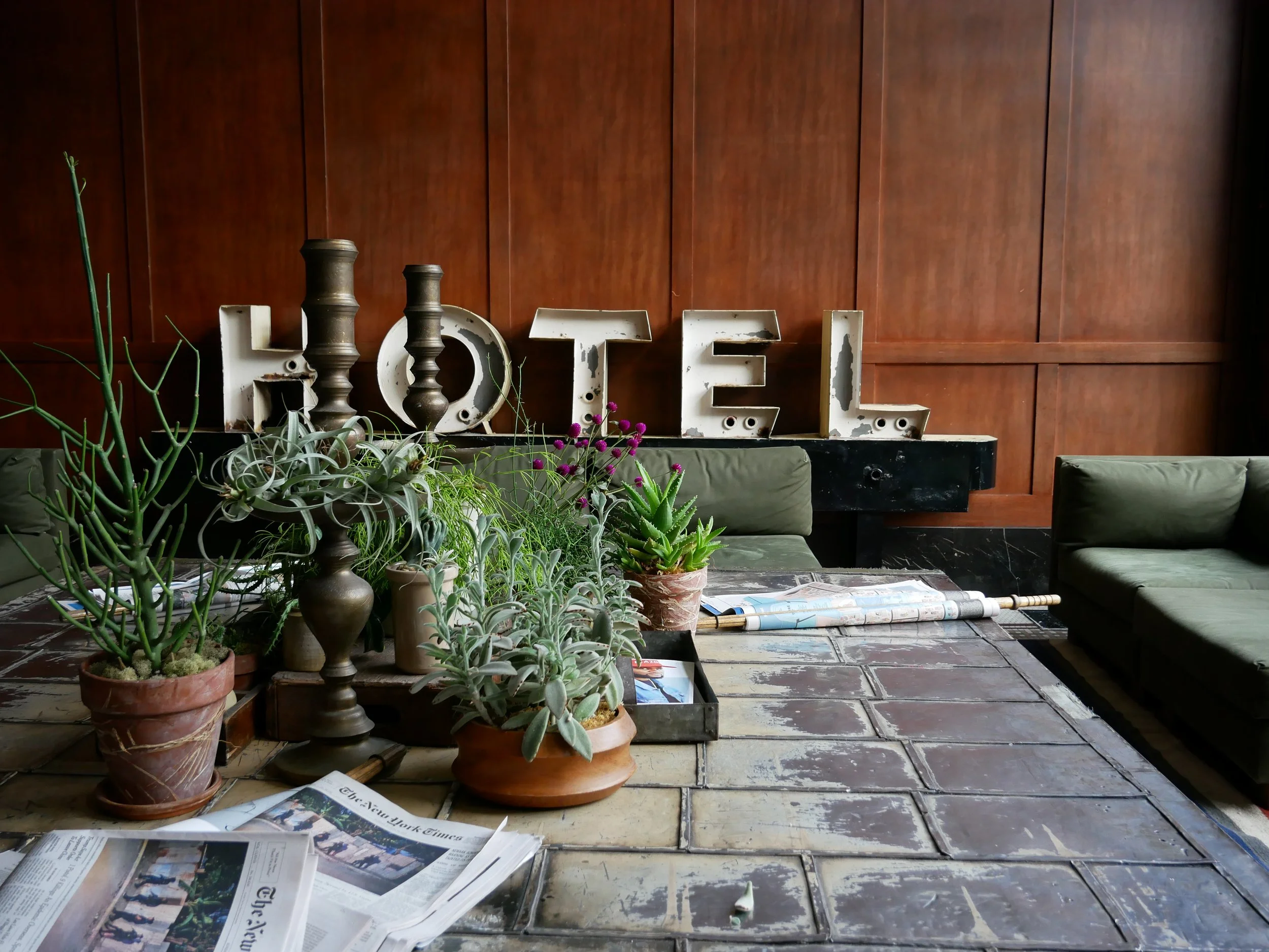 Une table en pierre décorée avec des pots de plantes grasses, un vase en bois et un magazine. Derrière, un mur en bois et des lettres en métal formant le mot 'HOTEL'.