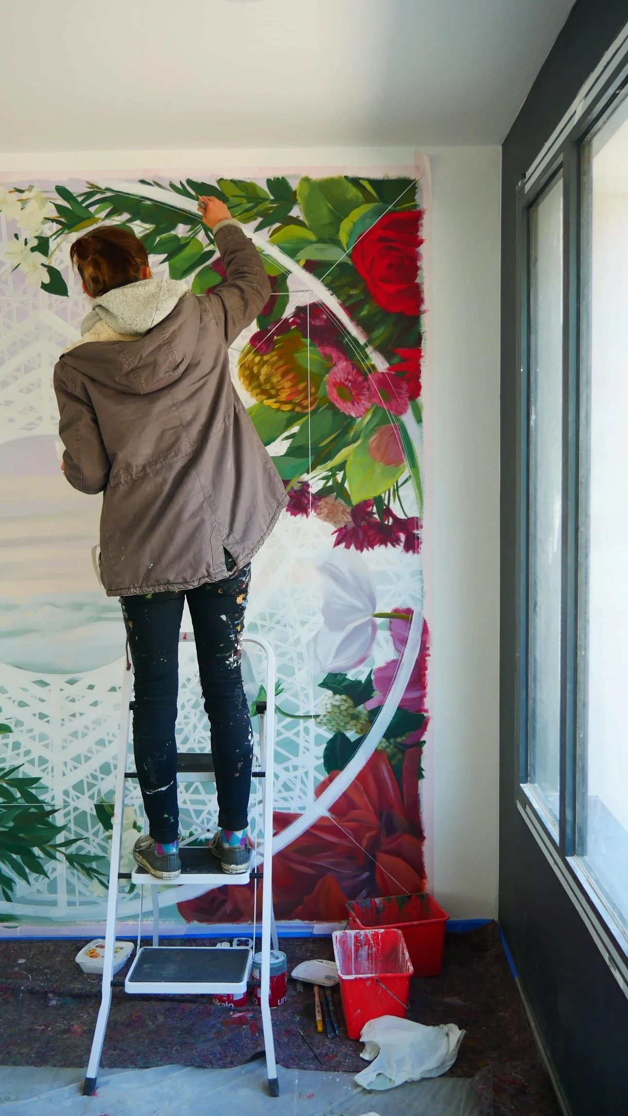 Une artiste peignant un mural floral coloré dans une pièce avec une fenêtre, utilisant une échelle pour atteindre le haut du mur. Réalisation de fresque pour la charte 1 immeuble 1 oeuvre