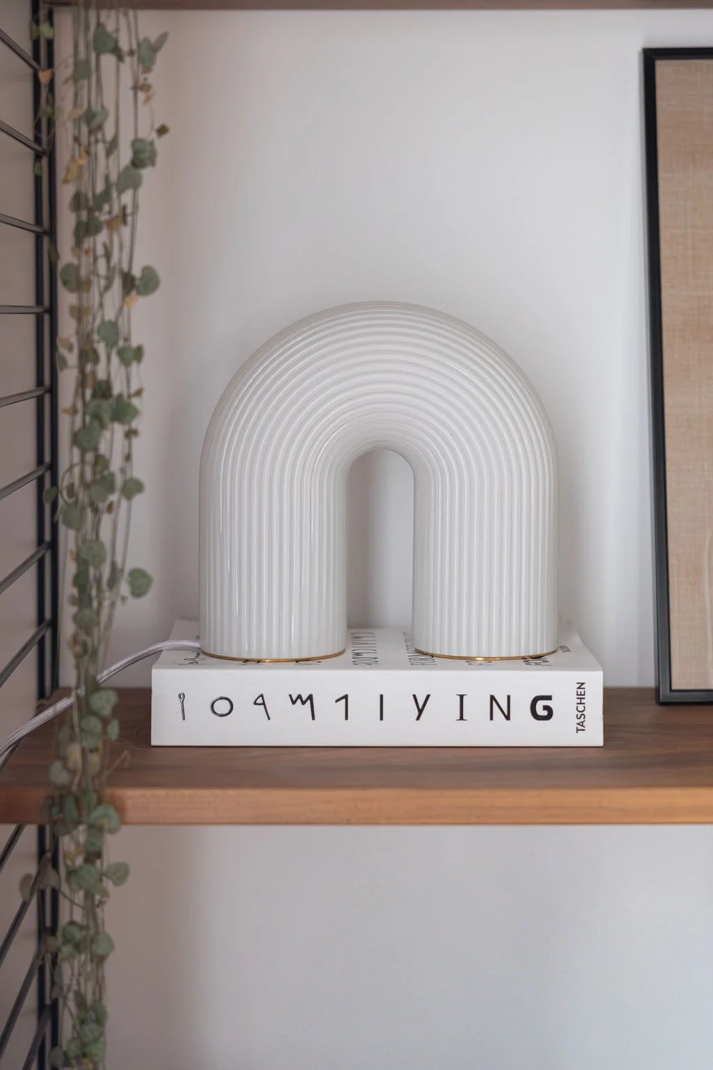 Lampe blanche en forme d'arc modèle Vuelta de la marque Ferm Living posée sur un livre blanc sur une étagère en bois, avec un cadre noir et un mur blanc en arrière-plan.