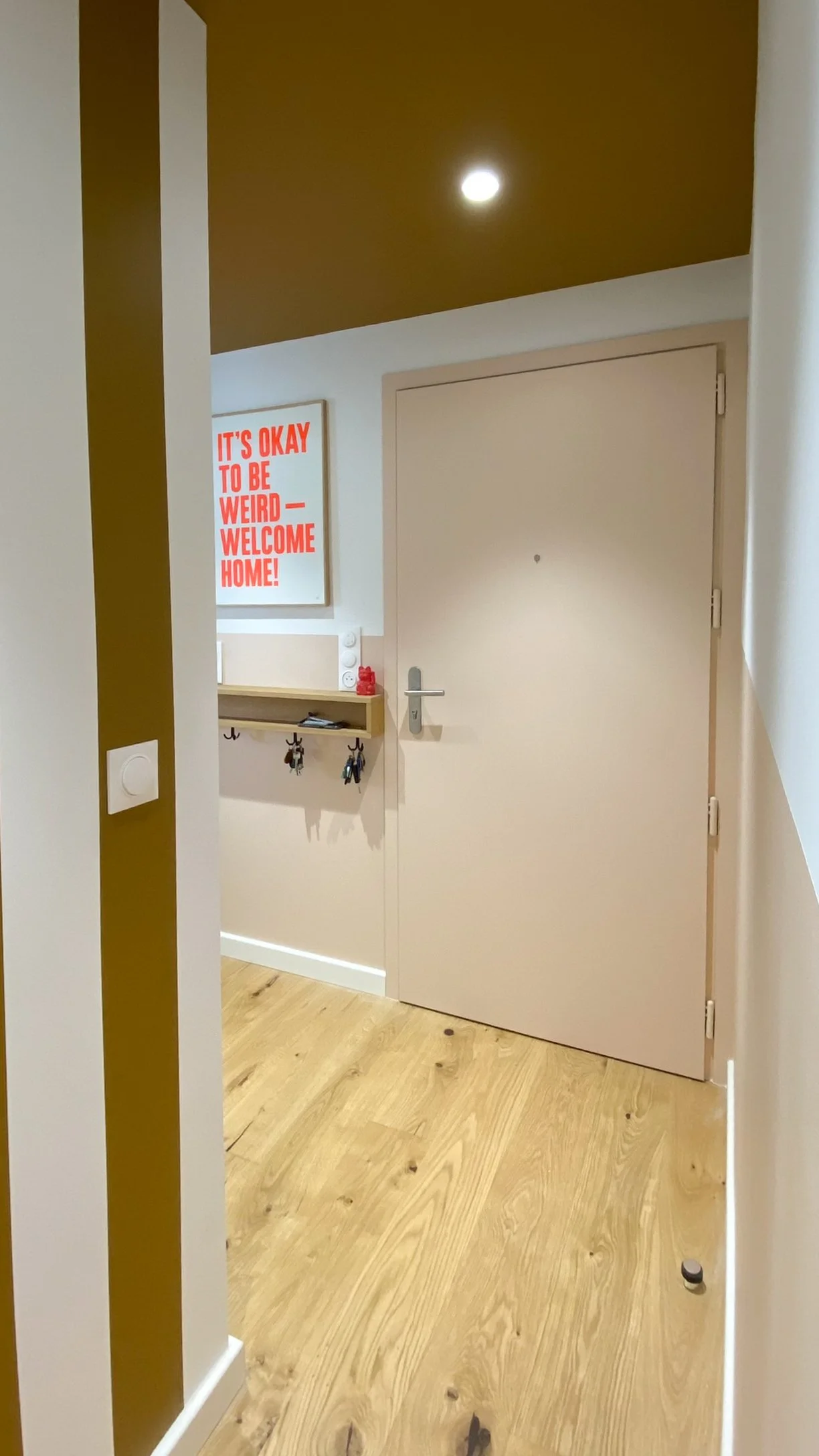 Vue d'une entrée avec une porte beige, un tableau avec le message en anglais 'It's okay to be weird - Welcome home', un porte-clés avec des clés et un meuble bois clair, sur le mur blanc.