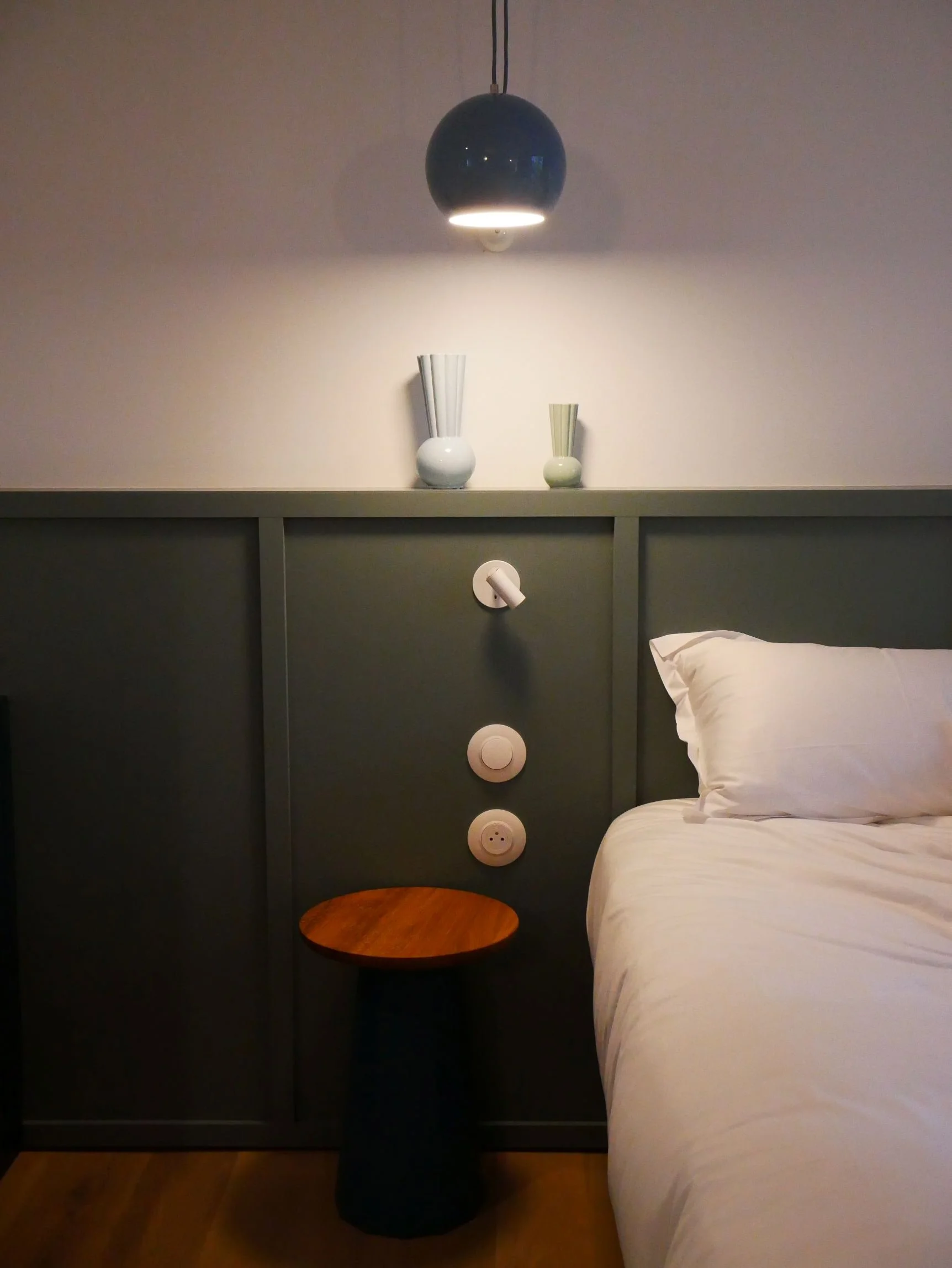 Chambre avec lit blanc, tête de lit gris foncé, lampe de chevet blanche, deux vases blancs avec des fleurs au mur, petit meuble en bois rond, prises électriques encastrées dans la tête de lit