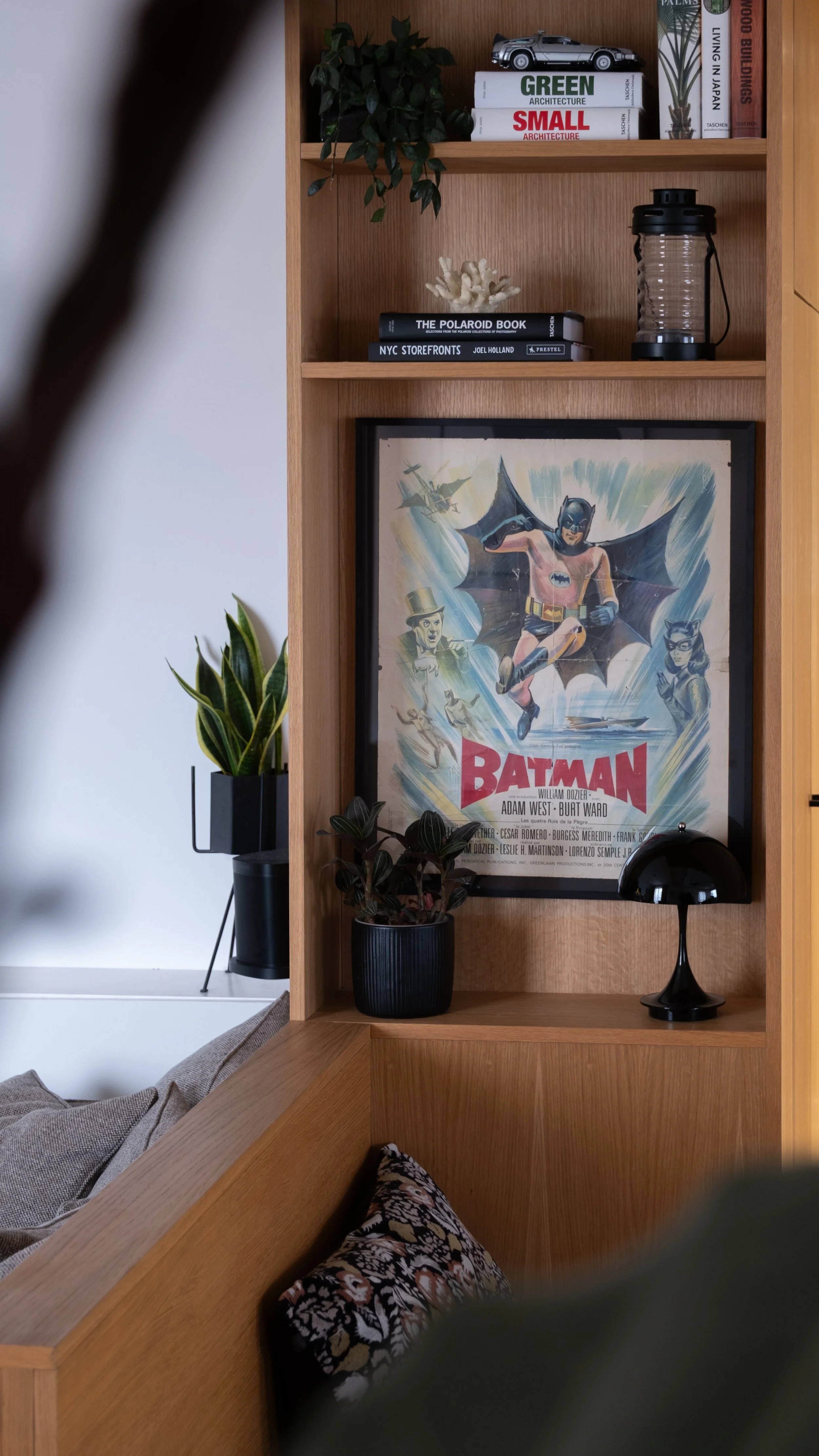 Étagère en bois avec des livres, une plante verte, une peluche de corail, un cadre avec une affiche vintage de Batman, une lampe noire modèle Panthella 160 de Louis Poulsen, et une petite lanterne.