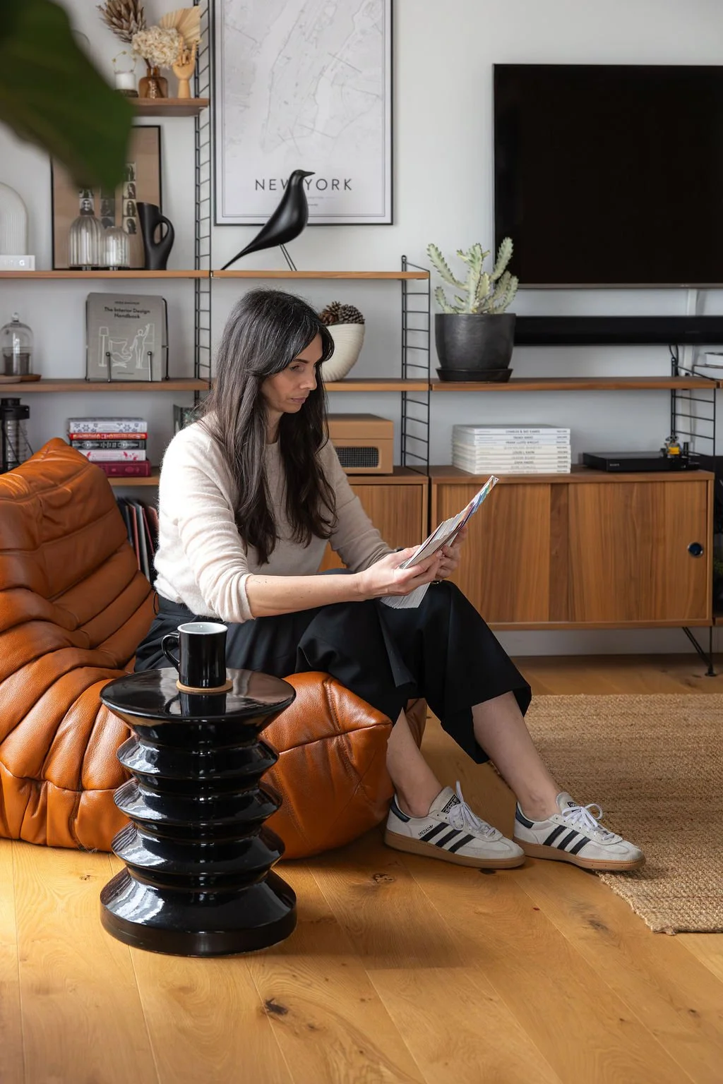 Femme assise sur un canapé en cuir marron, lisant un magazine, avec un tapis en jute à côté et un meuble en bois en arrière-plan, décoré avec des plantes, des livres et une télévision