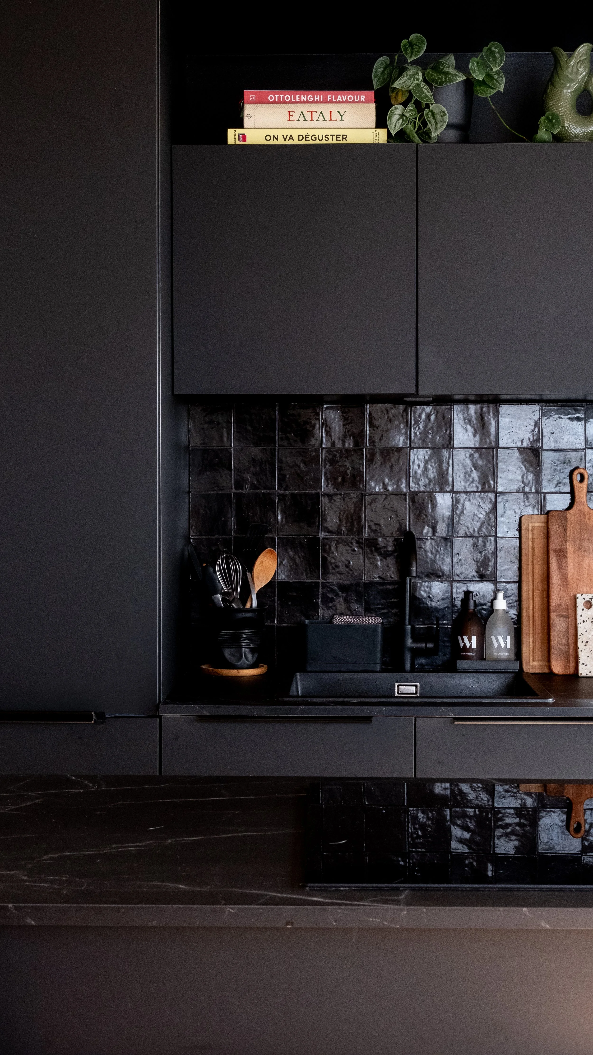 Cuisine noire avec plan de travail en marbre réalisée par Schmidt, échantillons d'ustensiles de cuisine dans un récipient noir, bouteilles en verre opale, planche à découper en bois, et étagère avec livres de cuisine et une plante verte.