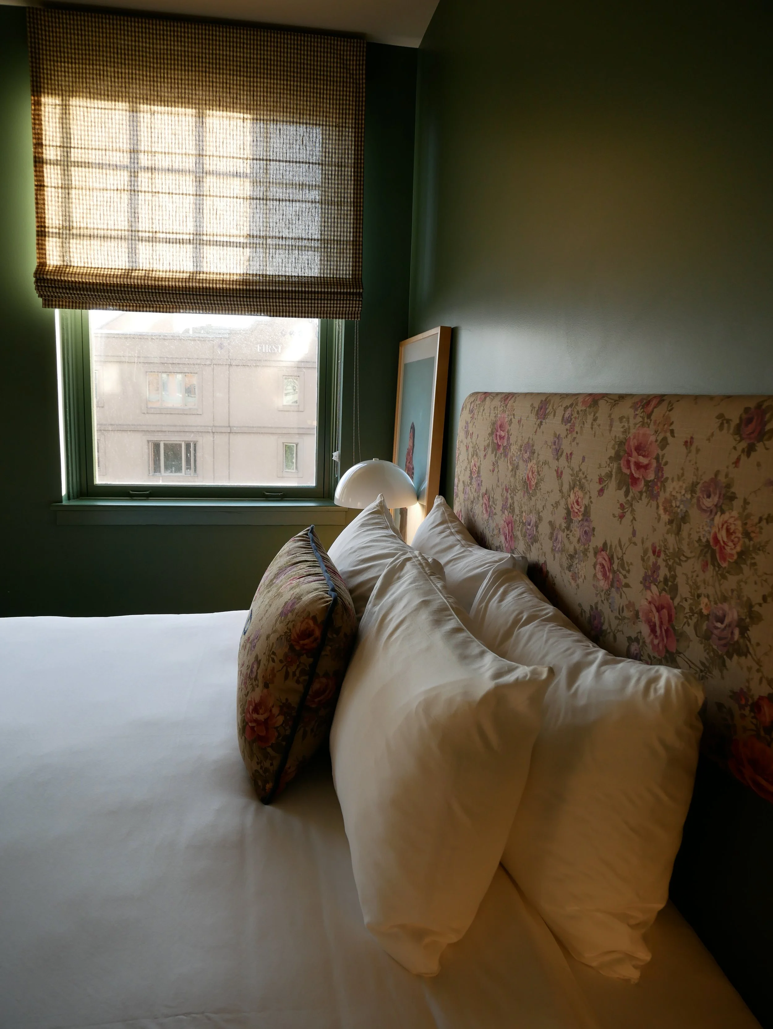 Lit avec des oreillers à motifs floraux, près d'une fenêtre avec un rideau à carreaux, dans une chambre à mur vert foncé.